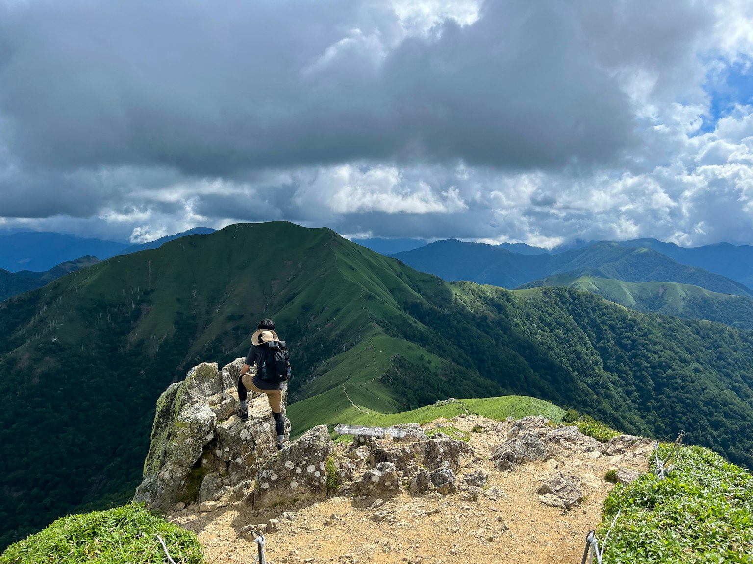 A hiker standing on a rock at the mountain summit with lush green hills in the background