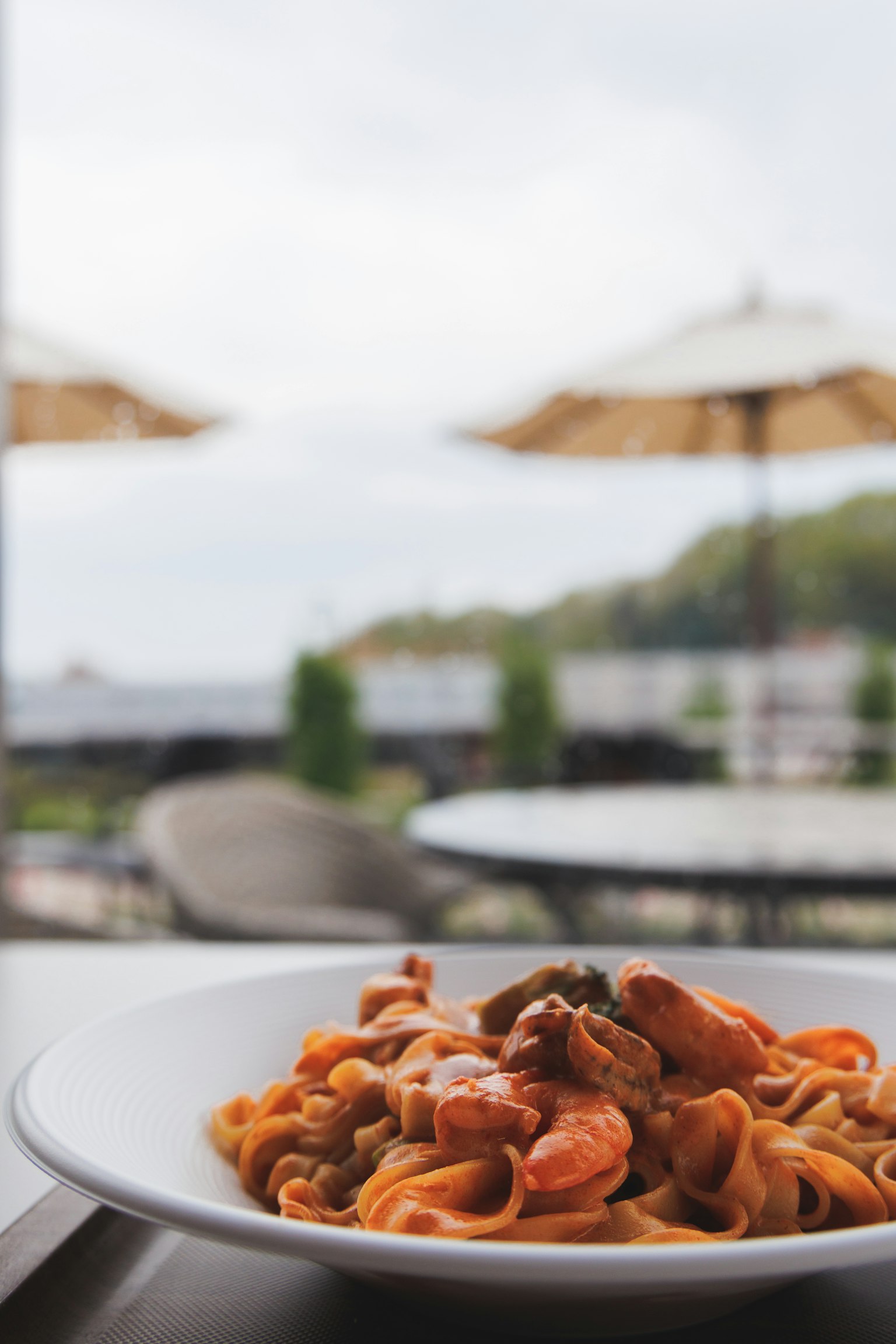 A plate of pasta with seafood in the foreground featuring tables with umbrellas and a seaside view in the background