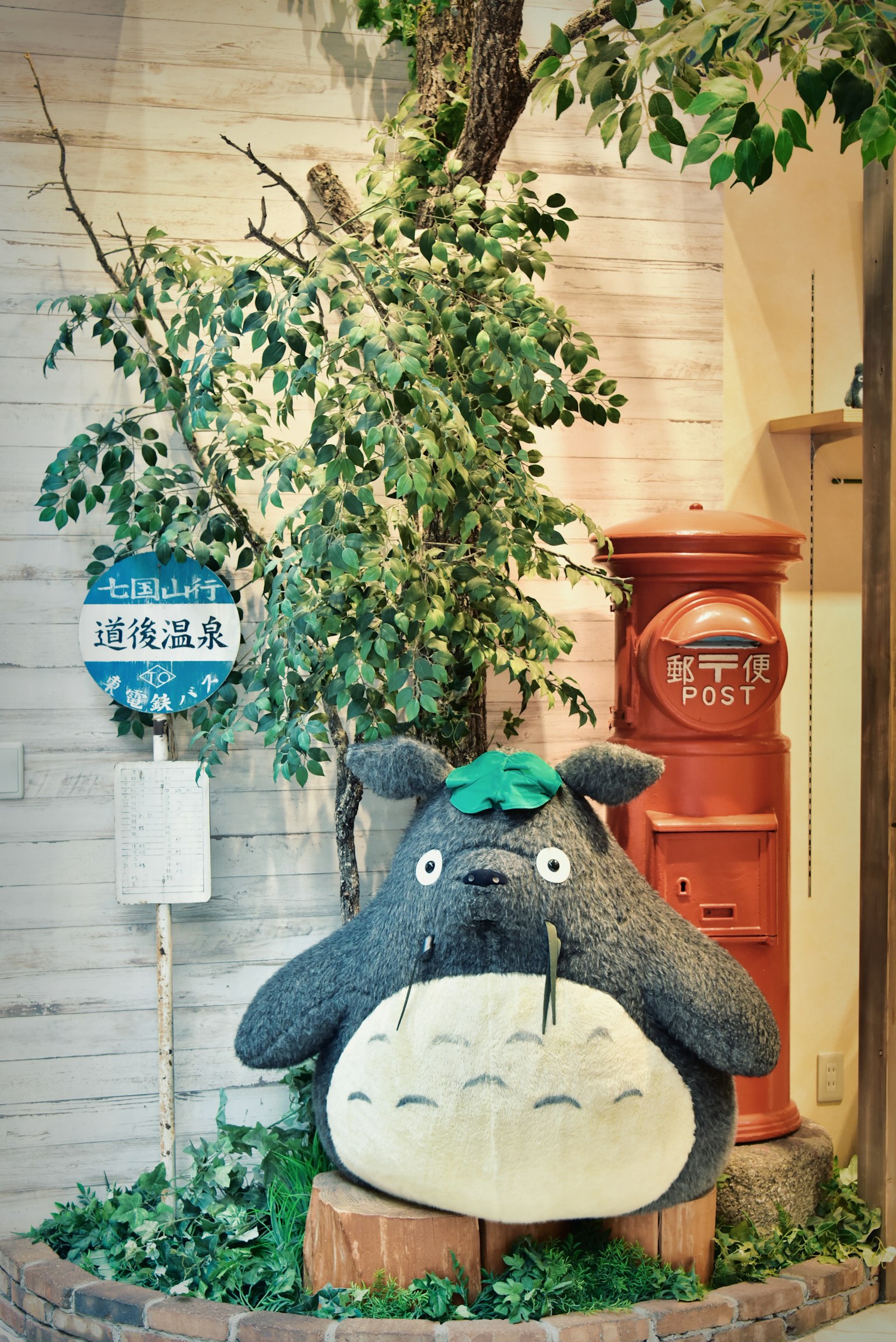 Un grand jouet en peluche Totoro entouré de verdure avec une boîte aux lettres rouge en arrière-plan