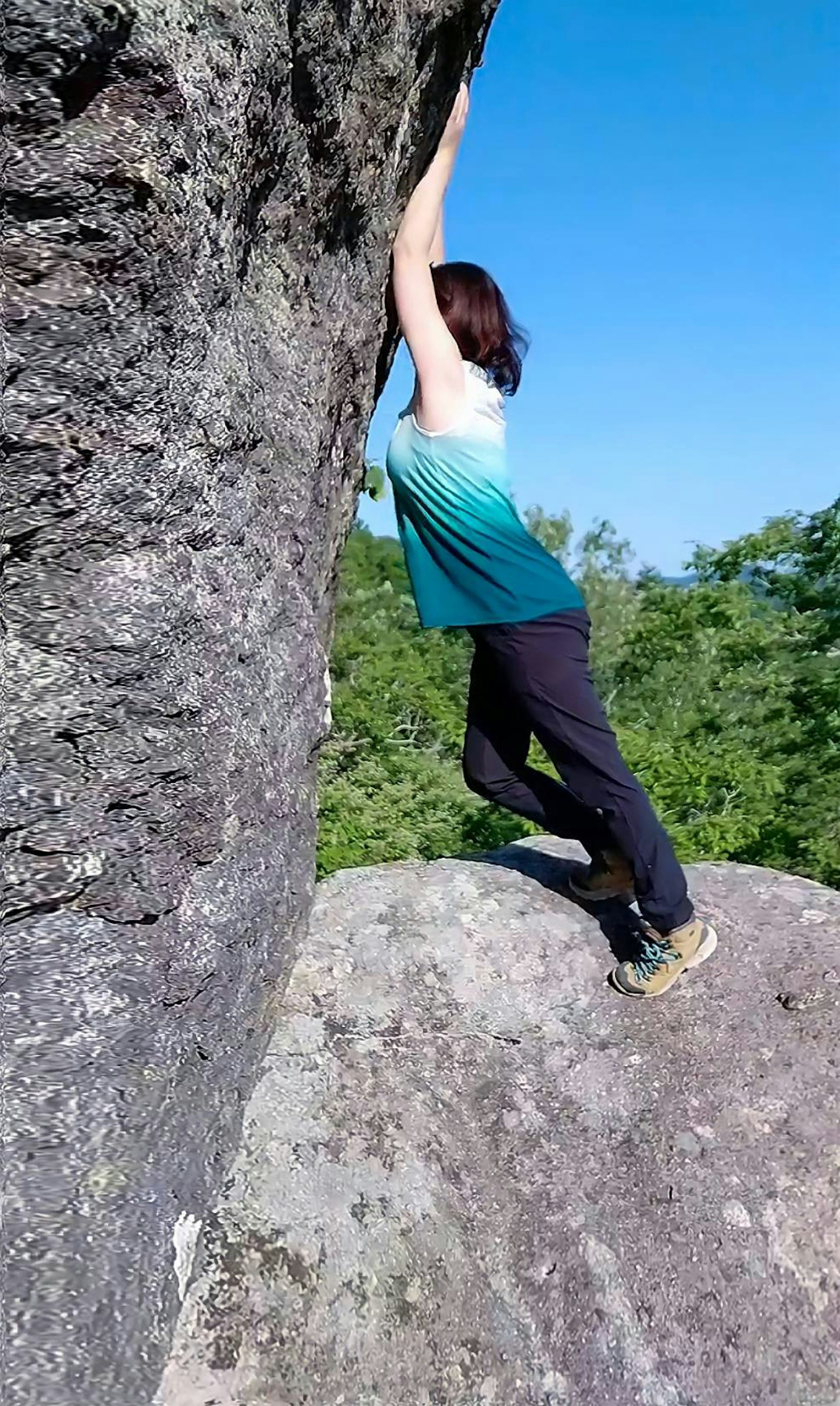 Woman climbing a rock wearing a green top and black pants under a blue sky