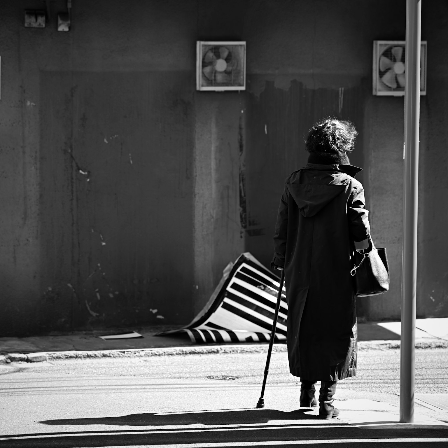 Una mujer caminando con un bastón en una escena de calle en blanco y negro con una tela de rayas en el fondo