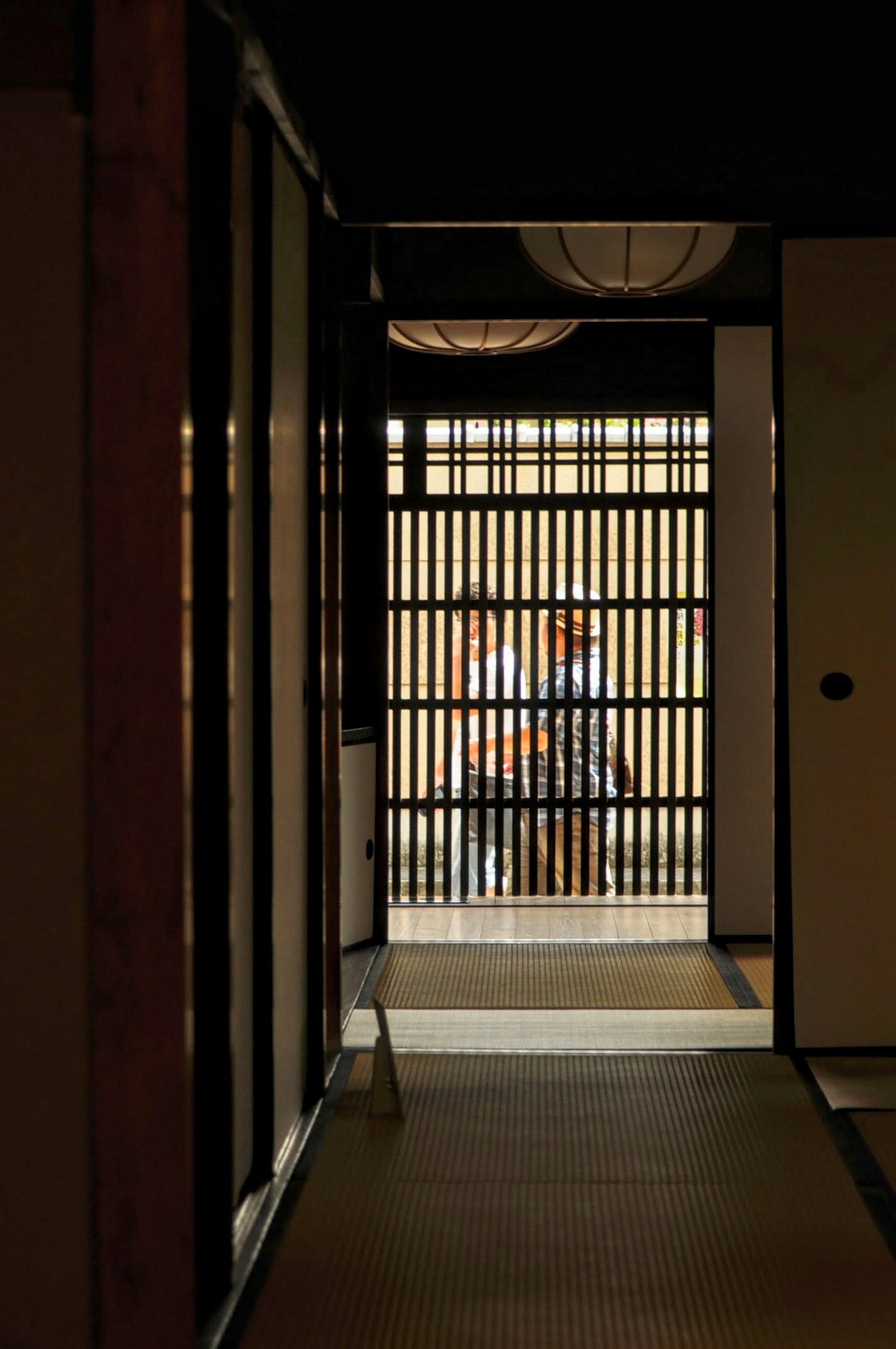 Blick durch einen dunklen Flur auf ein Gitterfenster und Menschen draußen