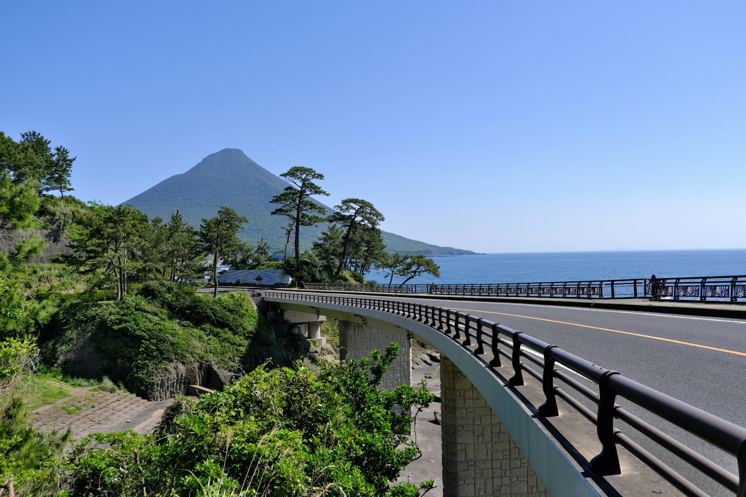 Jalan berkelok-kelok di tepi laut dengan latar belakang gunung di bawah langit biru yang jelas