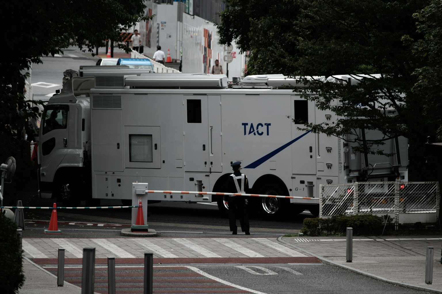 Un grand véhicule blanc marqué TACT dans une scène de rue avec un garde de sécurité surveillant la zone