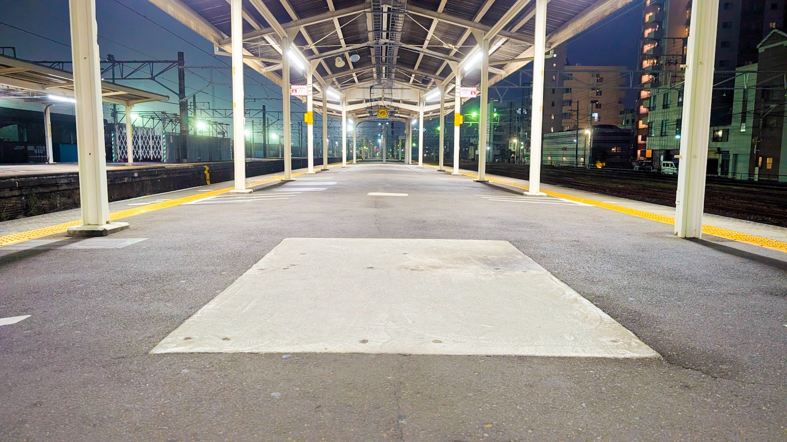 Plateforme de train vide la nuit avec des voies ferrées et des bâtiments environnants visibles