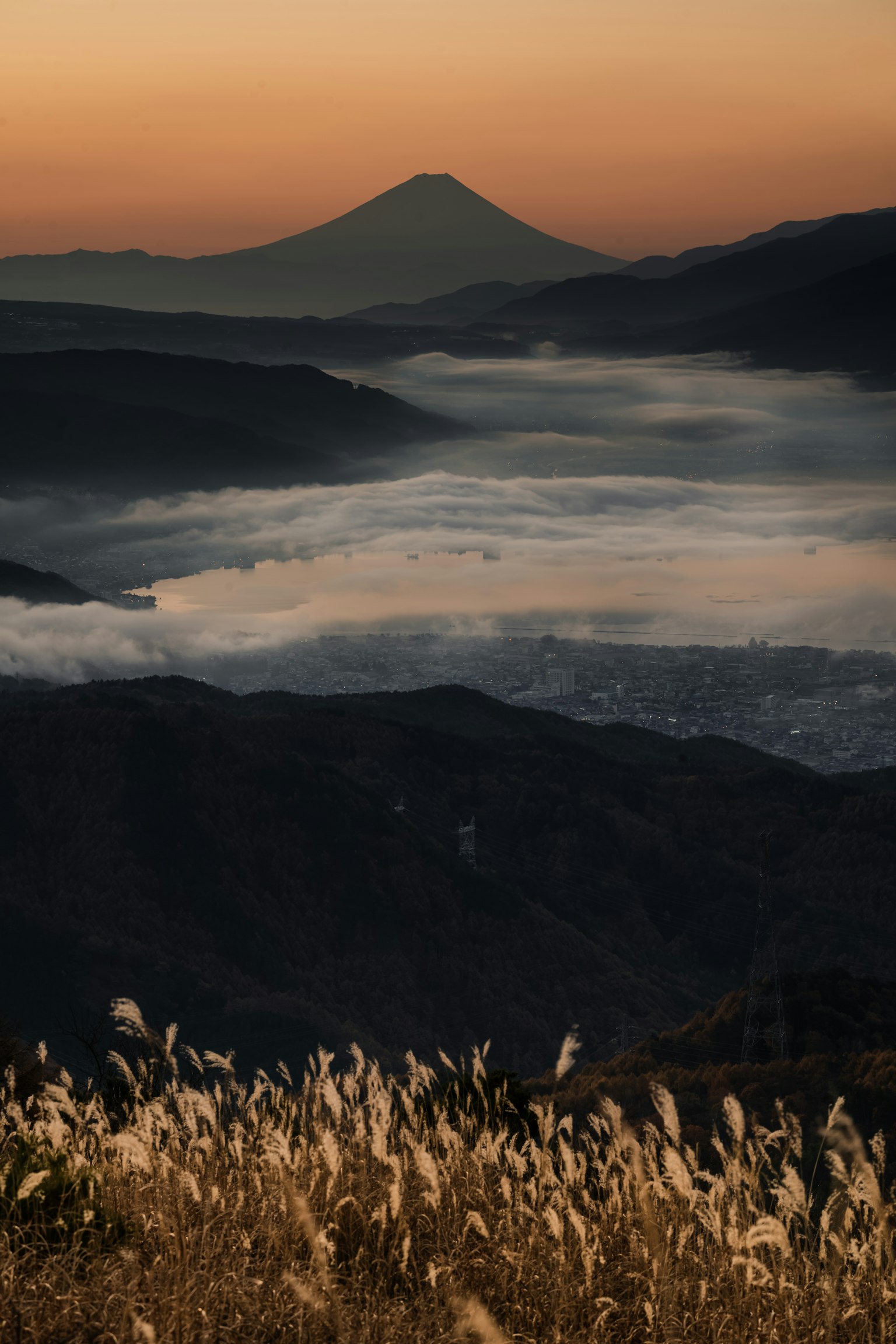 Vista escénica de niebla entre montañas con hierbas en primer plano silueta del Monte Fuji al atardecer