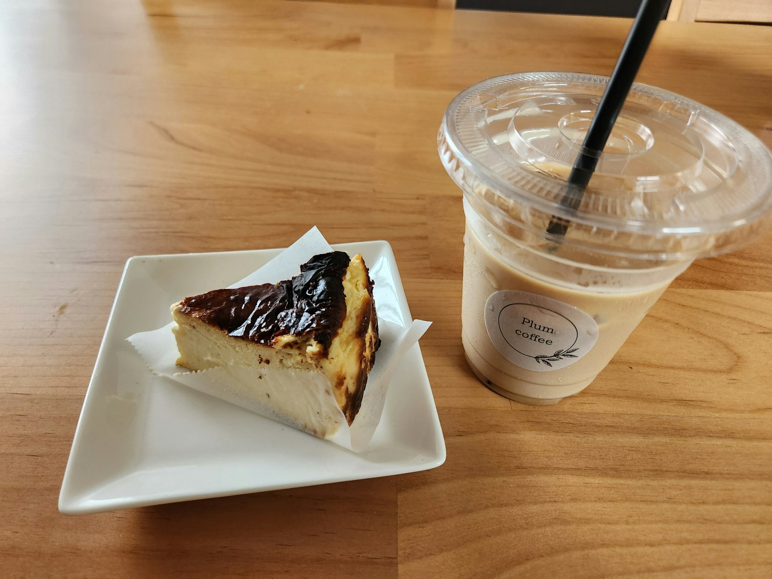 Ein Stück Käsekuchen auf einem weißen Teller neben einem Eiskaffee in einem durchsichtigen Becher