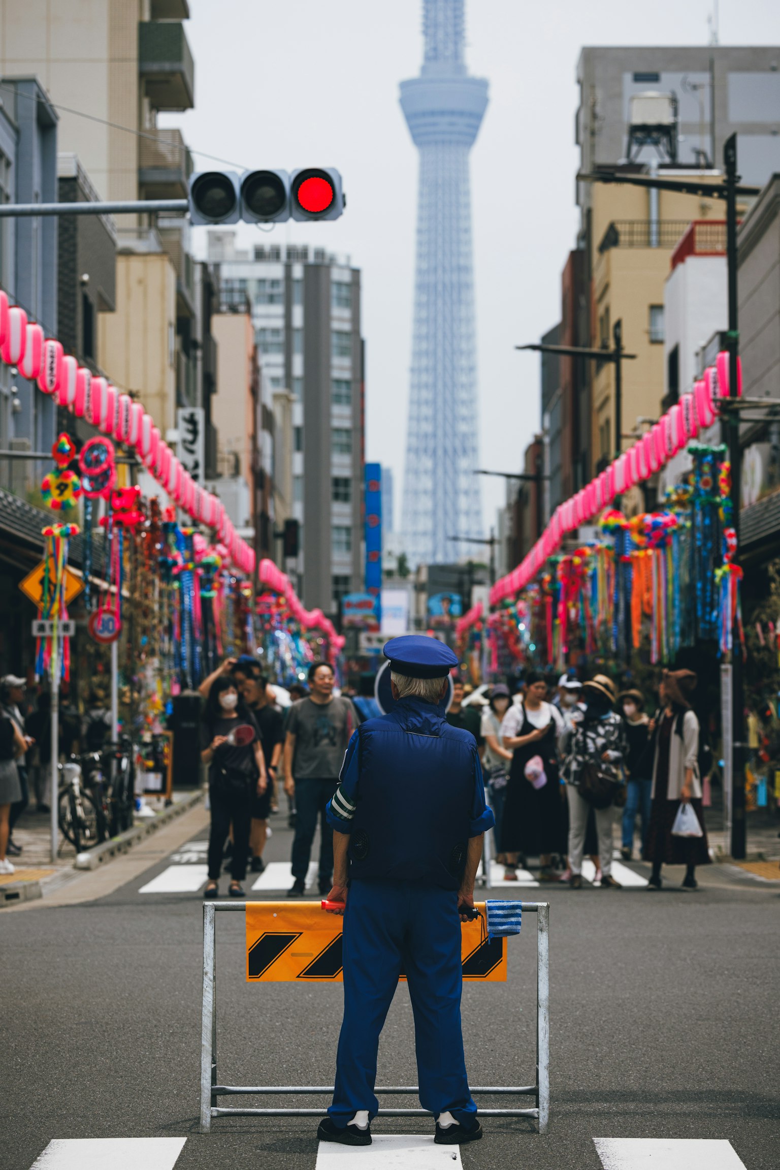Seorang polisi berdiri di depan penghalang di jalan yang ramai dengan lentera merah muda dan Tokyo Skytree di latar belakang