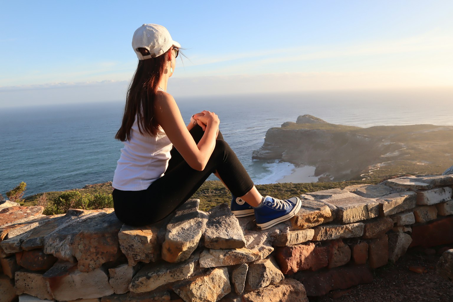 Donna seduta su un muro di pietra che guarda l'oceano indossando un cappellino bianco pantaloni neri e sneakers
