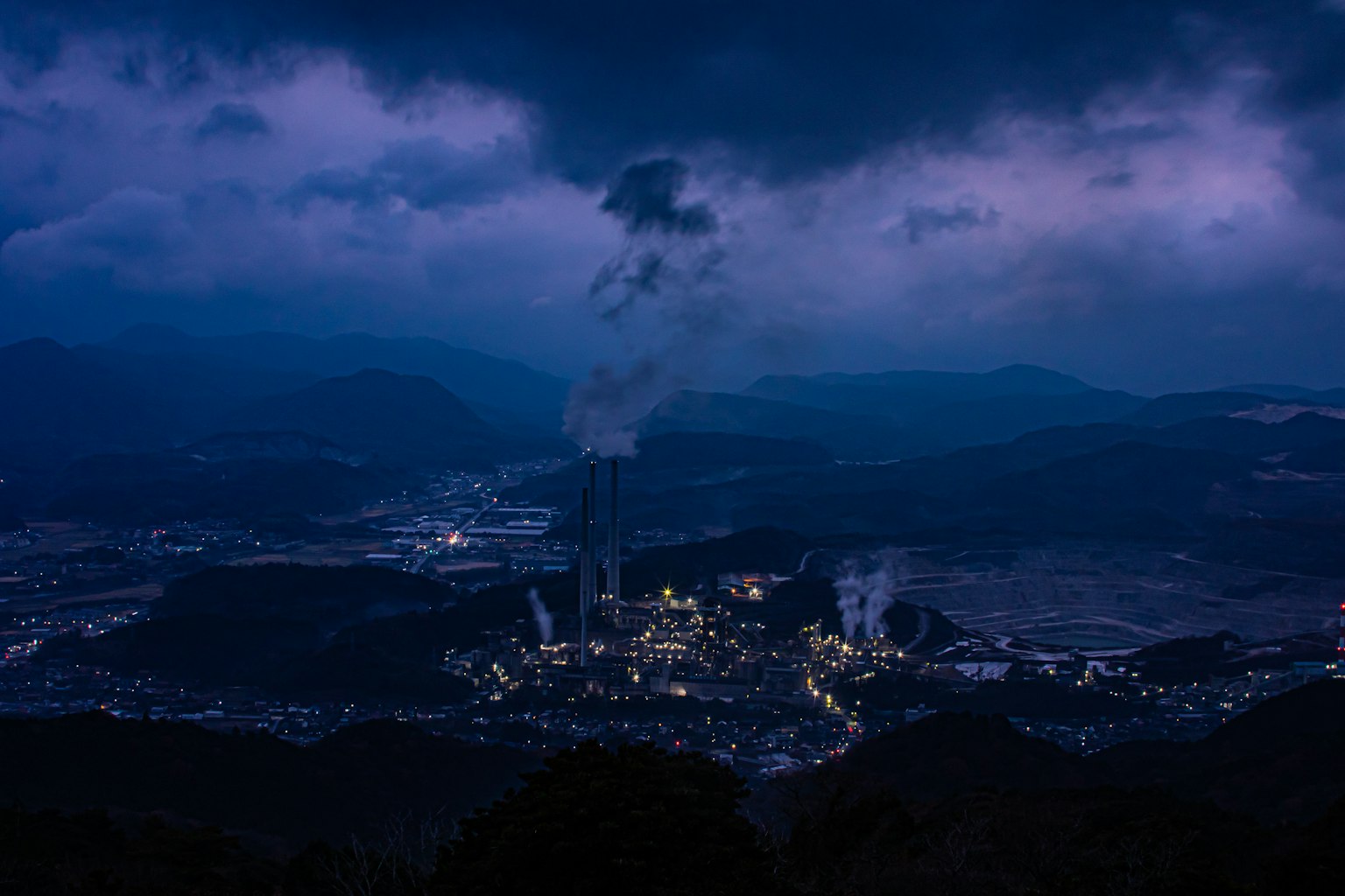 Vista nocturna de una zona industrial con chimeneas humeantes y montañas circundantes