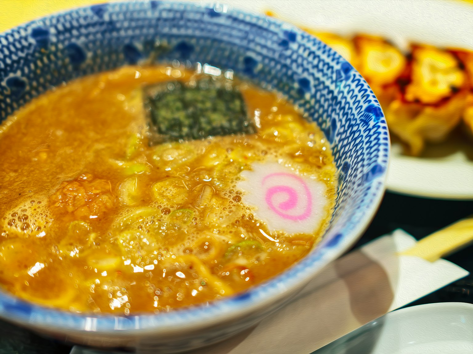 Ramen dalam mangkuk biru dengan hiasan rumput laut dan narutomaki di samping gyoza panggang