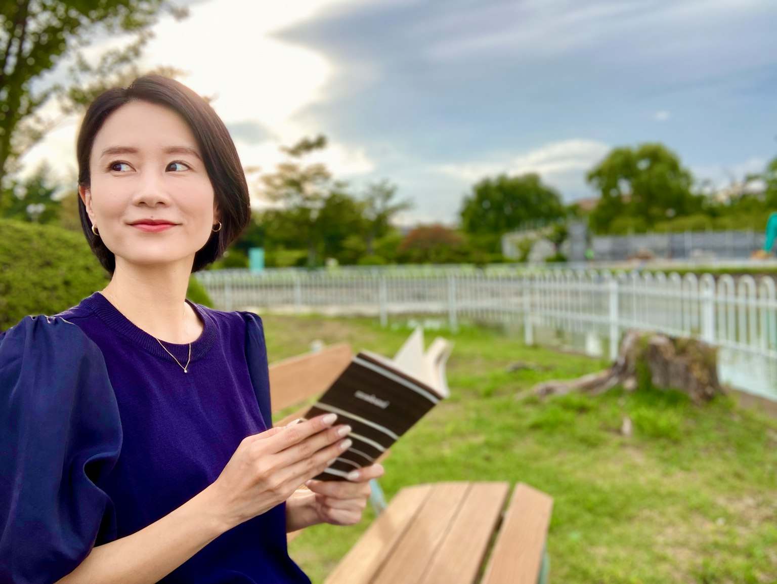 Potret seorang wanita tersenyum sambil memegang buku di taman
