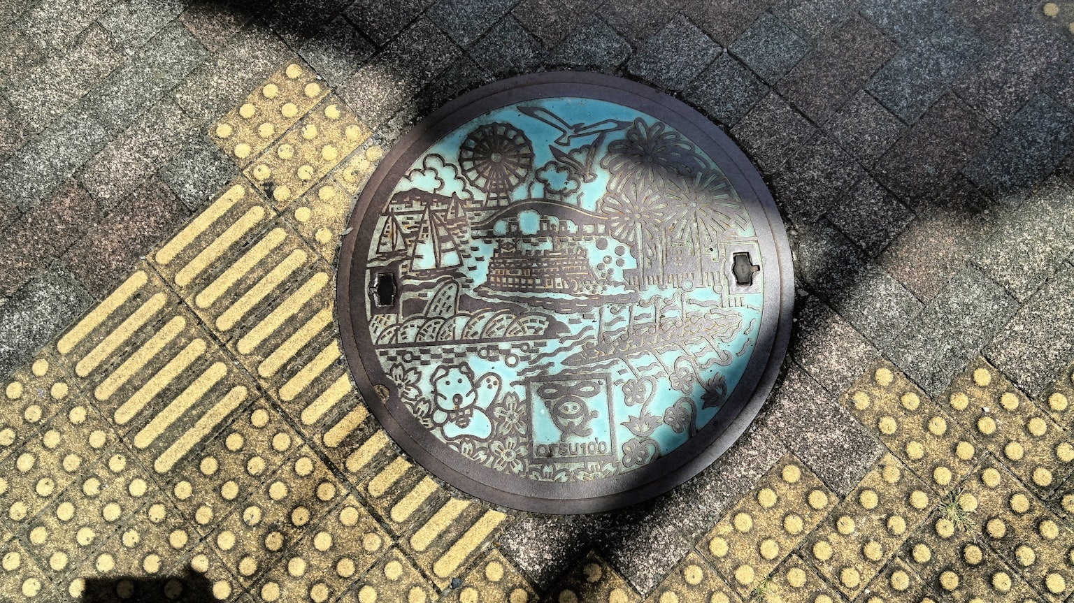 Coperchio di pozzetto blu con design e pavimentazione tattile gialla circostante