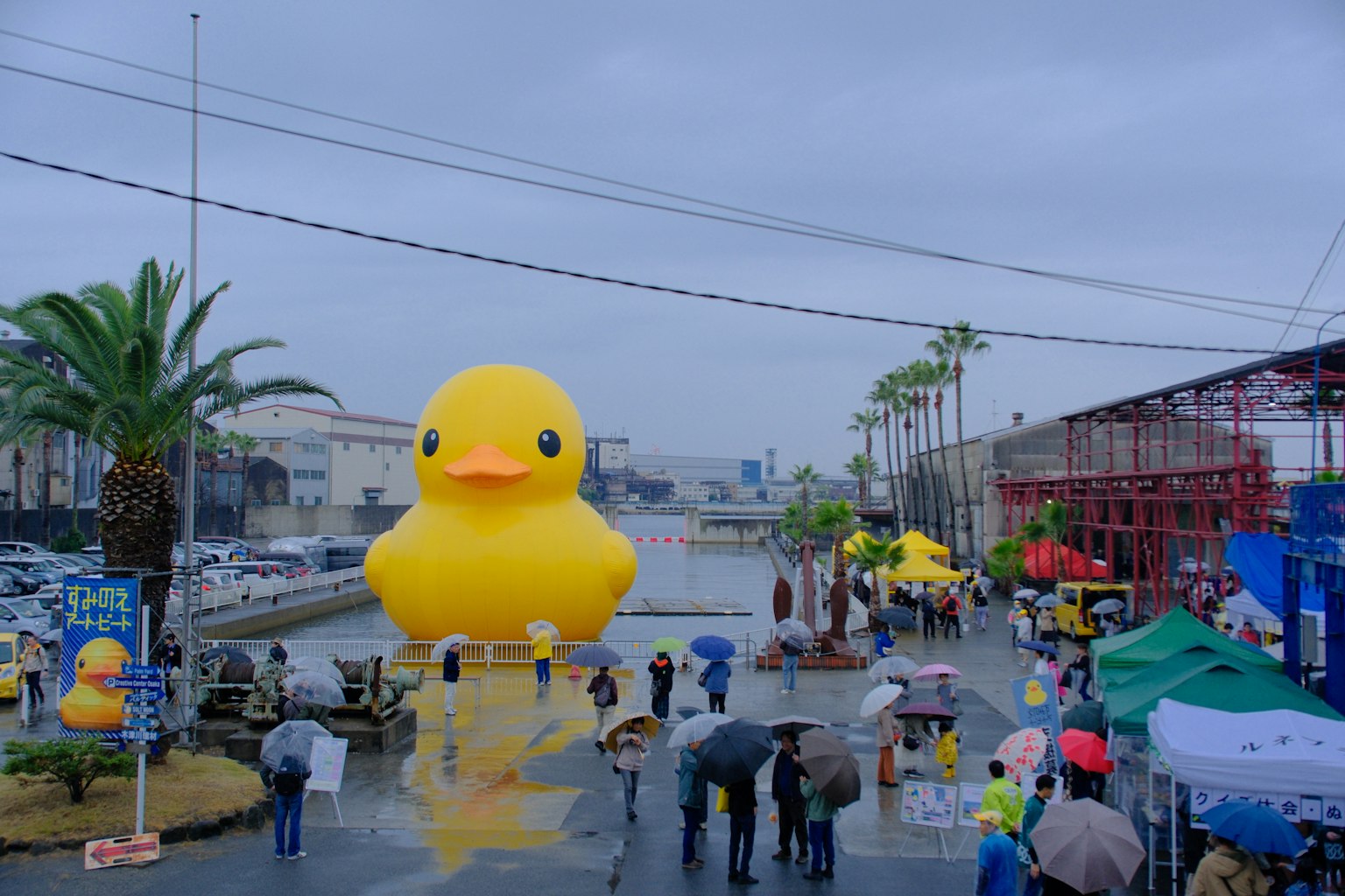 大きな黄色いアヒルのバルーンが水辺に浮かんでいる雨の日の風景