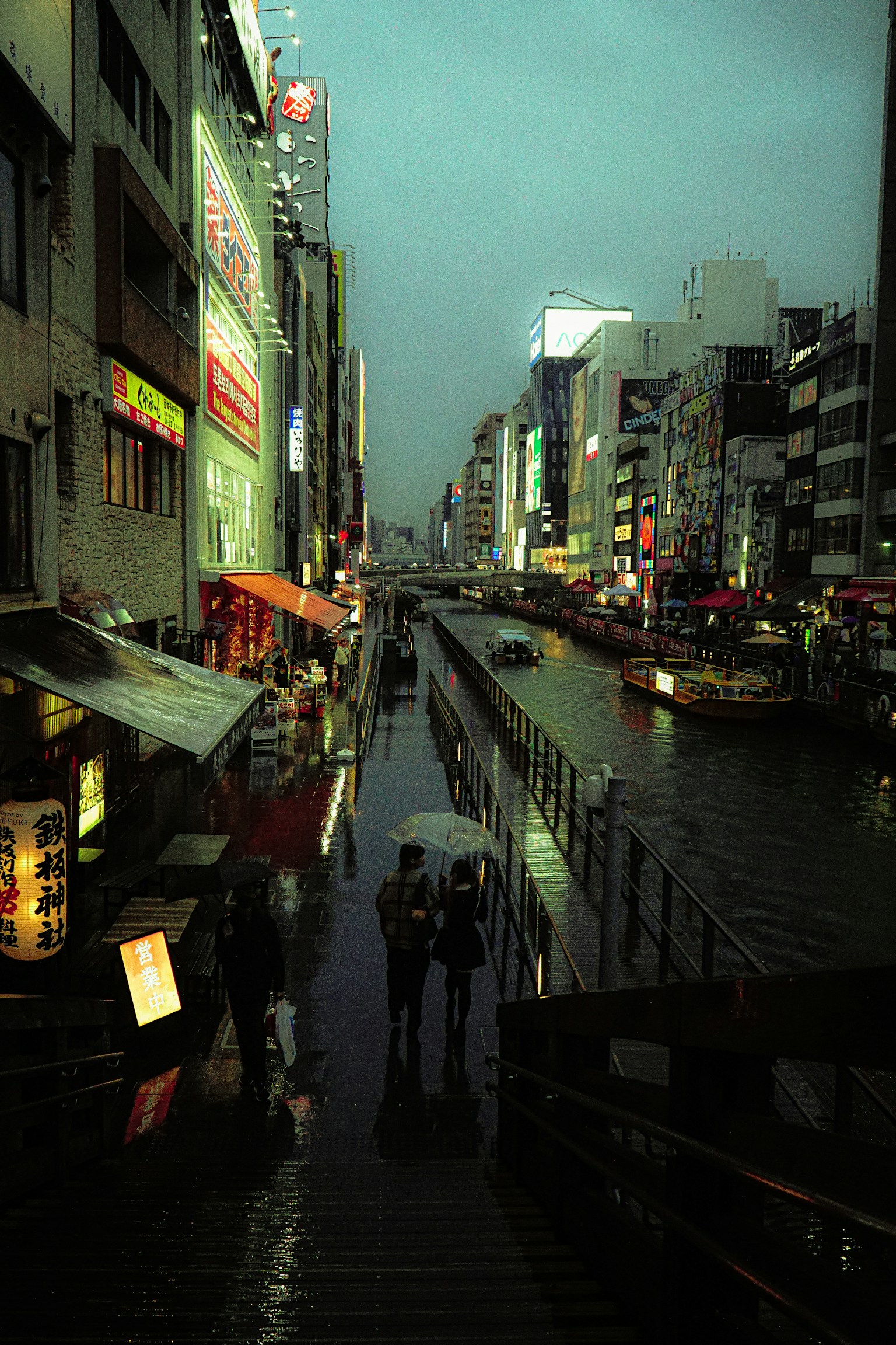 夜間城市景觀沿河而建，燈光閃爍的商店和人們的輪廓