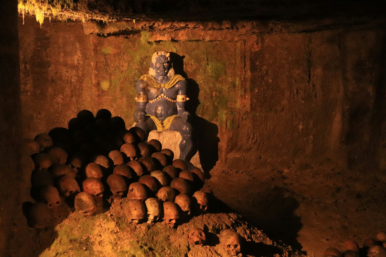 Eine blaue Statue sitzt in einem unterirdischen Raum zwischen einem Haufen Schädel