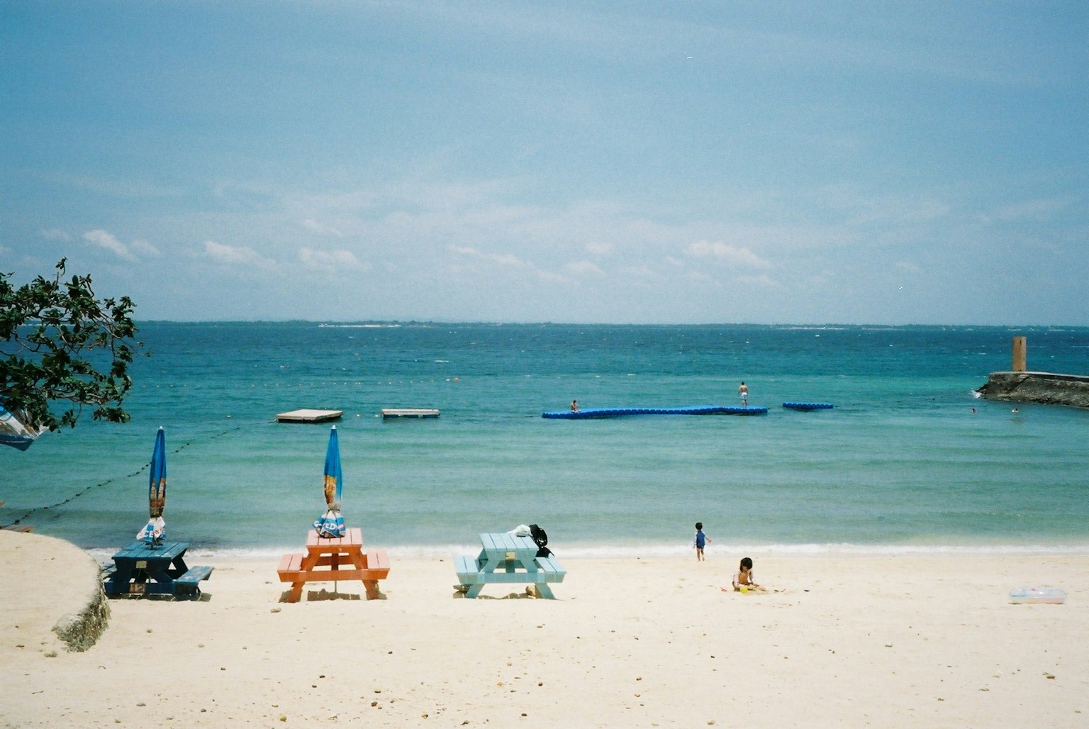 โต๊ะปิกนิกหลากสีสันบนชายหาดทรายกับทะเลสีน้ำเงินใสและเรือในระยะไกล