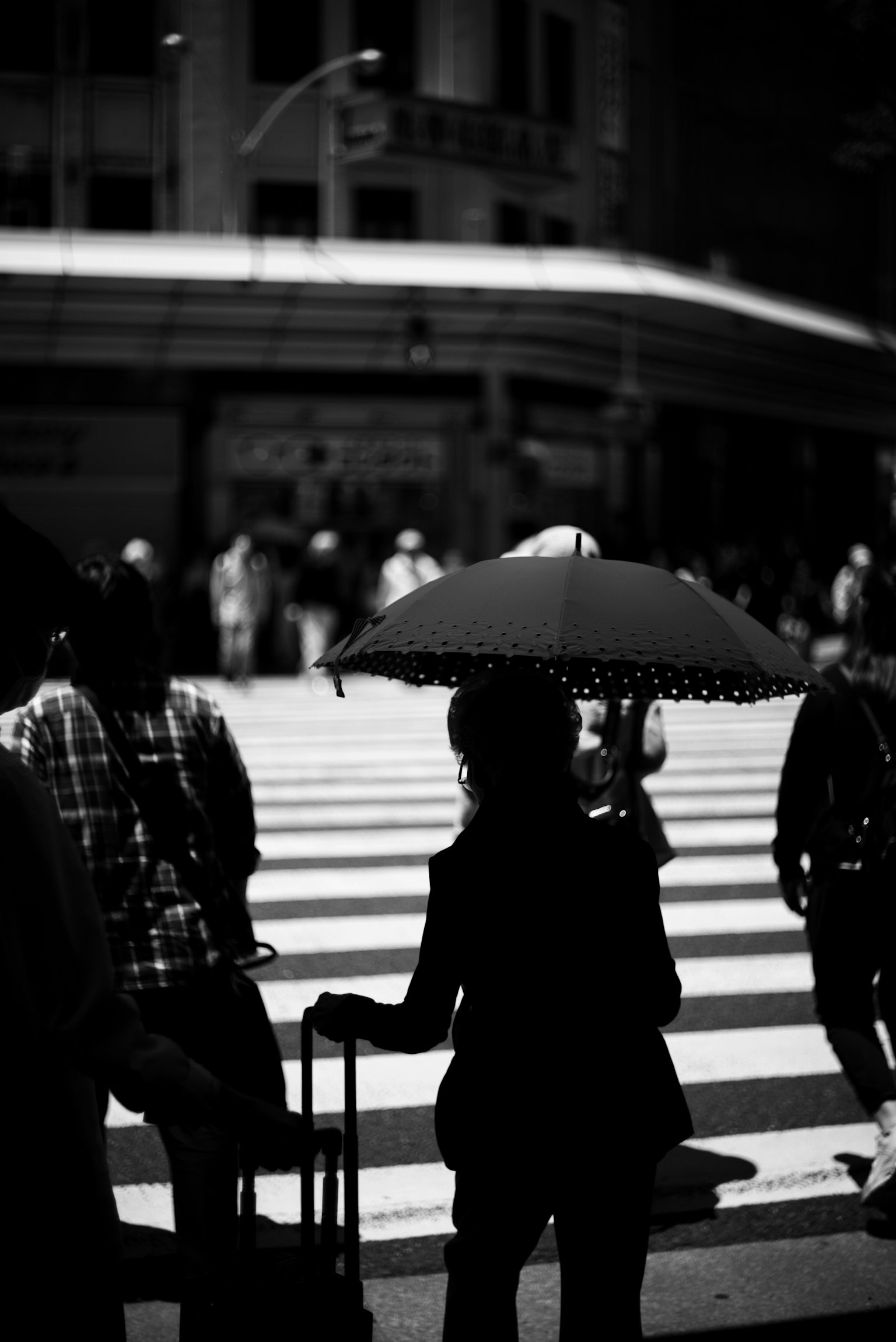 Silhouette of a woman with an umbrella standing at a crosswalk surrounded by pedestrians in a black and white city scene