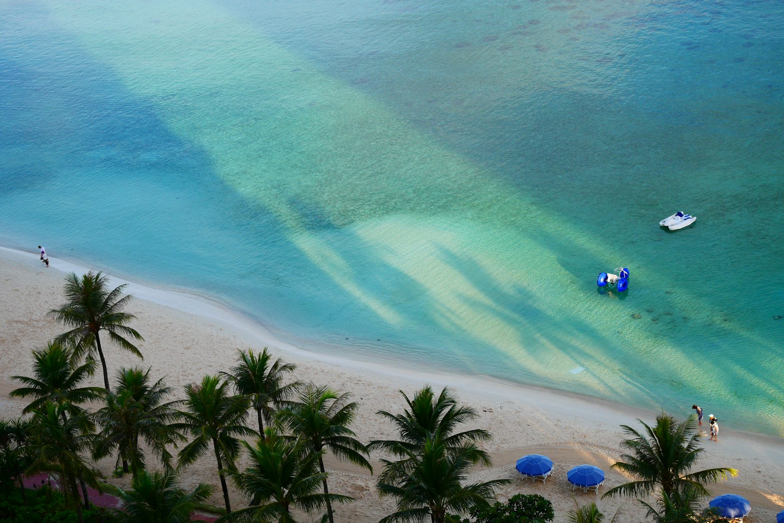 Aerial view of a beach with blue water and white sand featuring palm trees and blue umbrellas