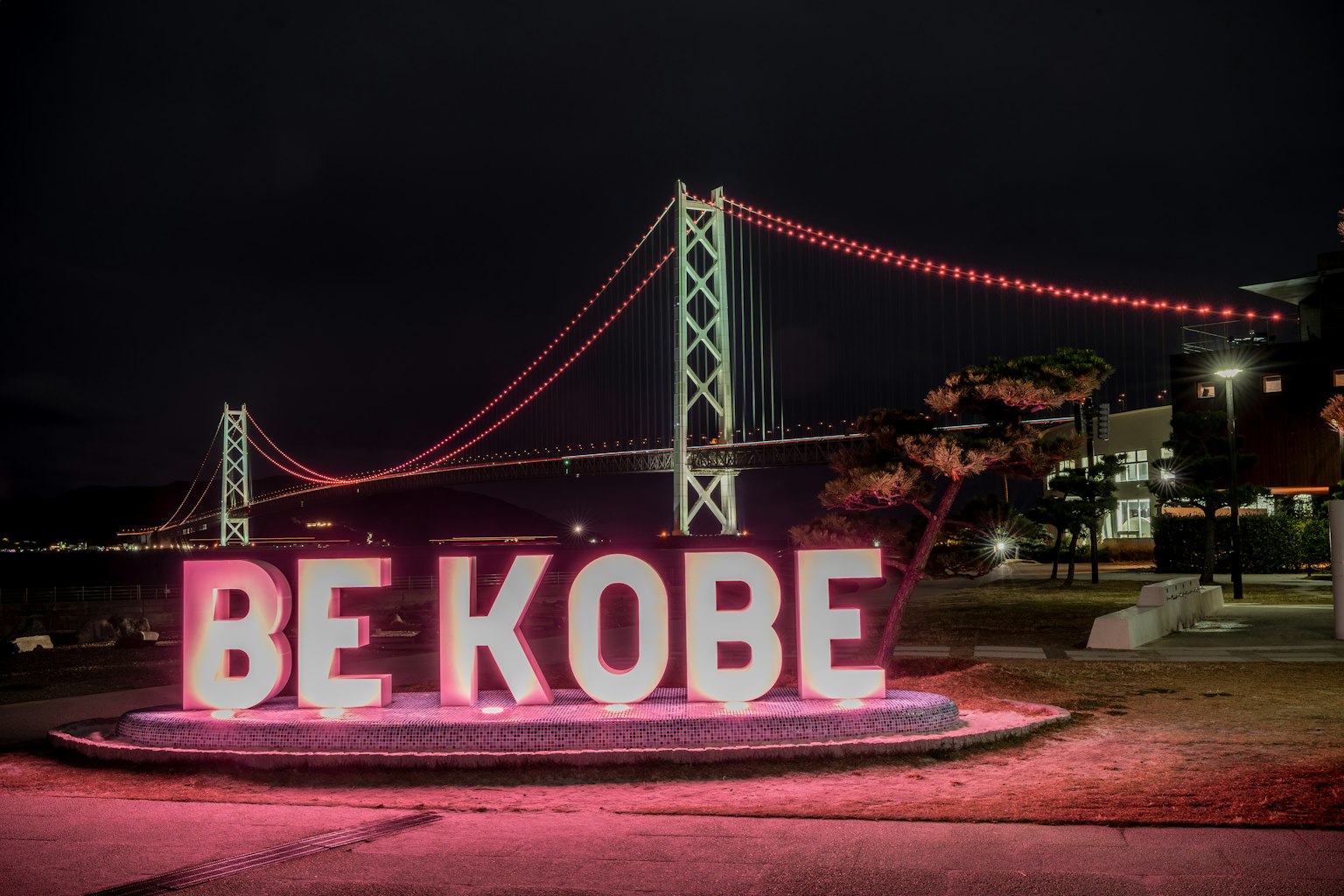 Grandi lettere illuminate che formano BE KOBE con un ponte decorativo sullo sfondo di notte