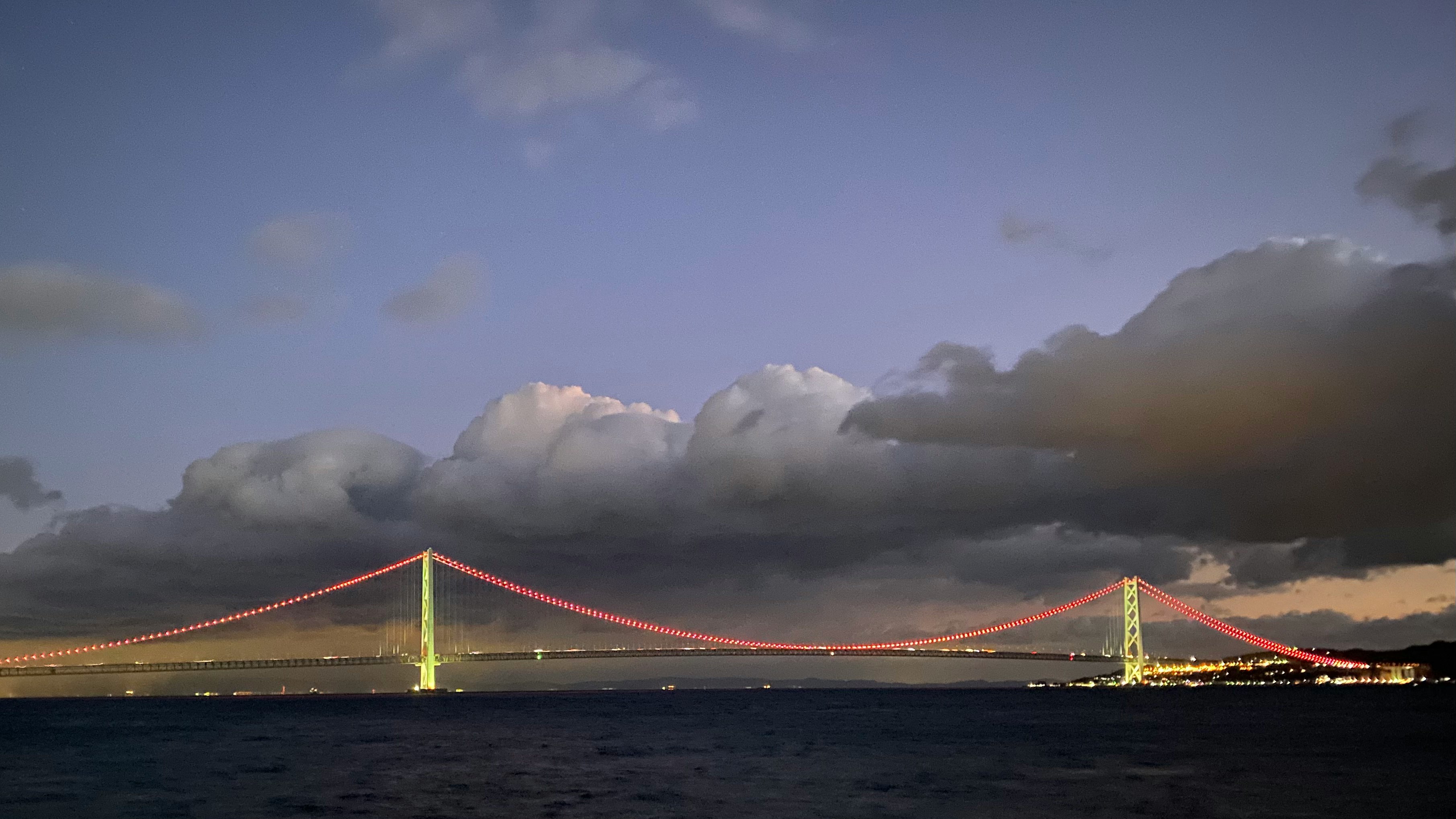 夕暮れの海上にかかる赤い吊り橋と雲の空