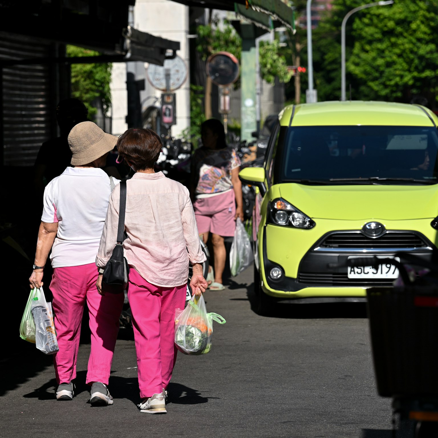 Dua wanita mengenakan celana pink berjalan dengan tas belanja di jalan kota Mobil hijau melintas di dekatnya