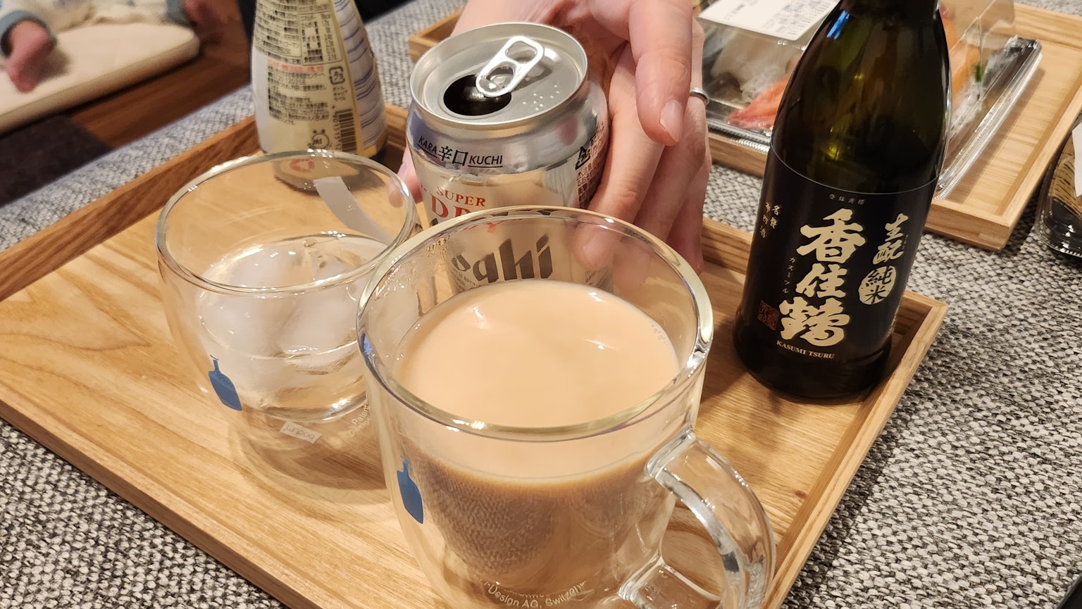 A set of beverages on a wooden tray with a transparent glass containing ice a mug filled with a brown drink and a black sake bottle