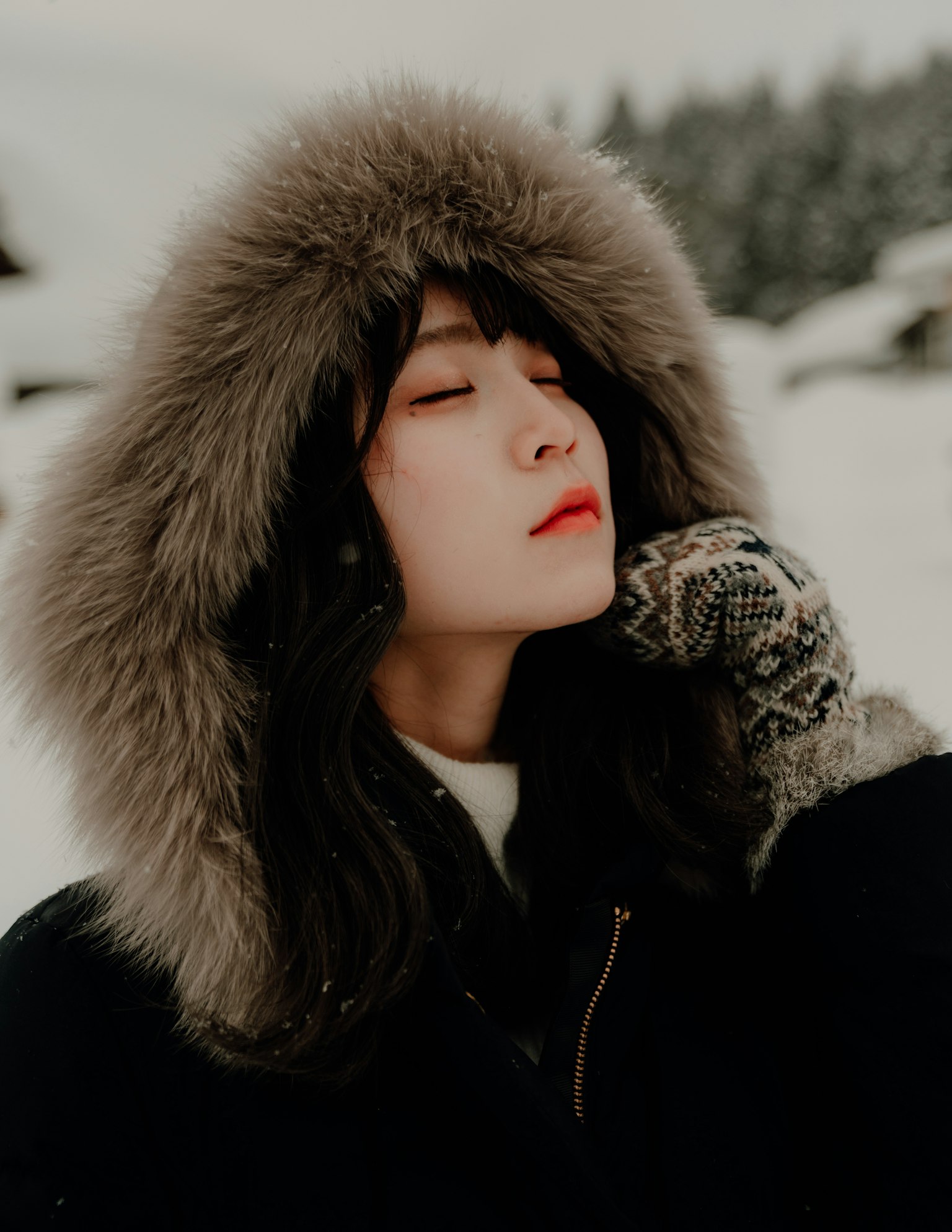 Woman with closed eyes in snow wearing a black coat with fur hood and gloves