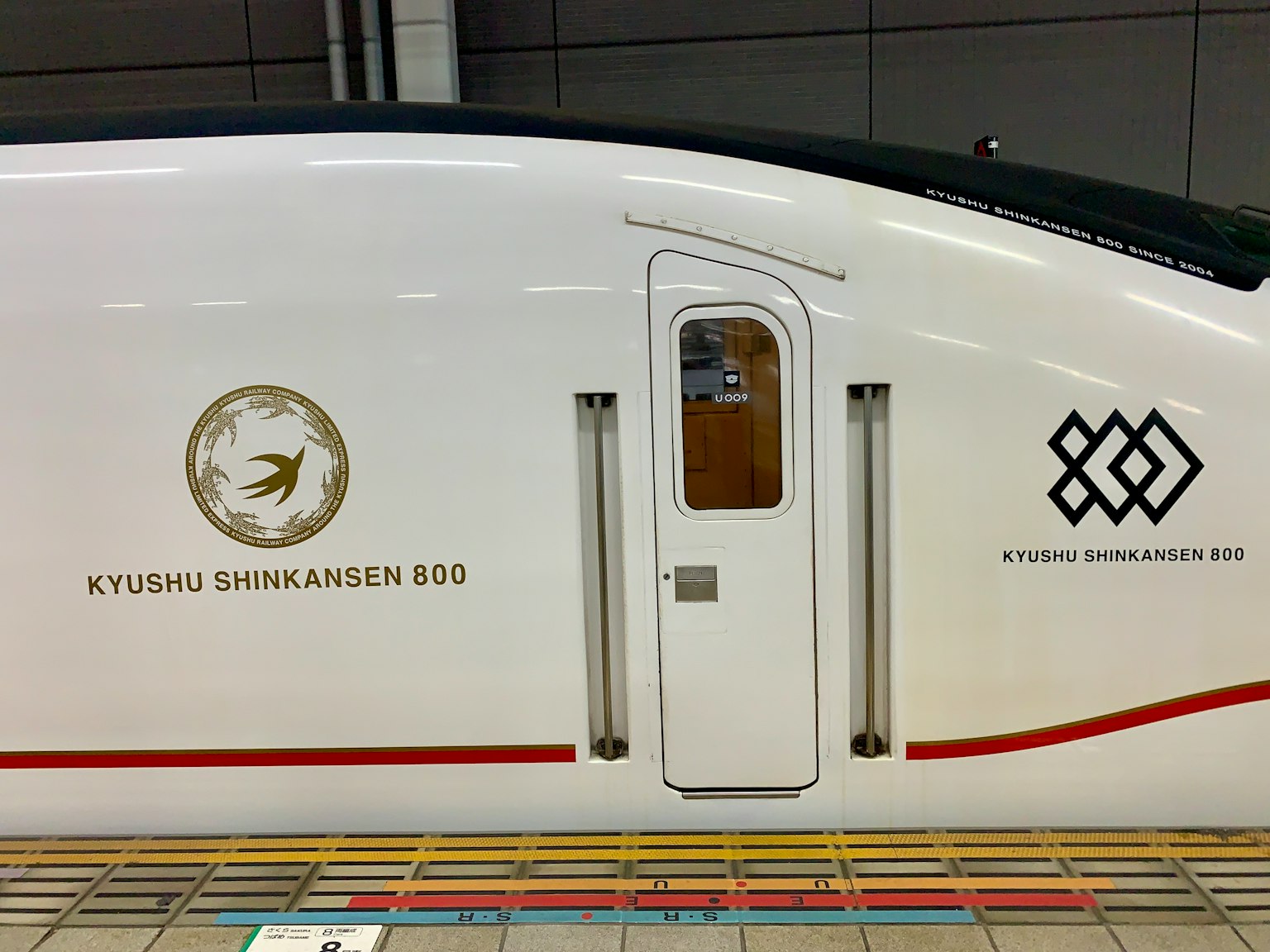 Tampilan samping kereta Shinkansen putih Kyushu dengan logo dan pintu