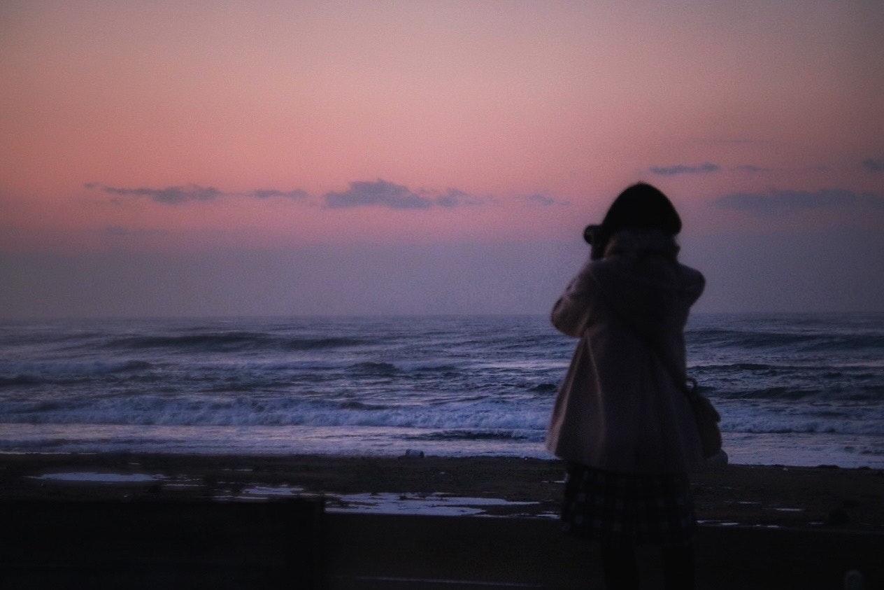 冬の日本海の浜辺で夕暮れに海を撮るカメラ女子の後ろ姿