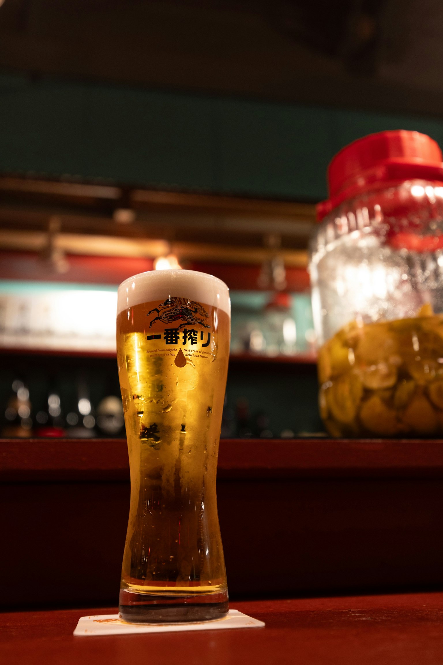 Un verre de bière sur un comptoir en bois avec un grand bocal en arrière-plan contenant des cornichons