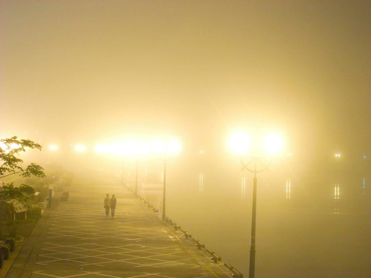 Zwei Figuren, die auf einem nebligen Weg am Fluss gehen, beleuchtet von Straßenlaternen