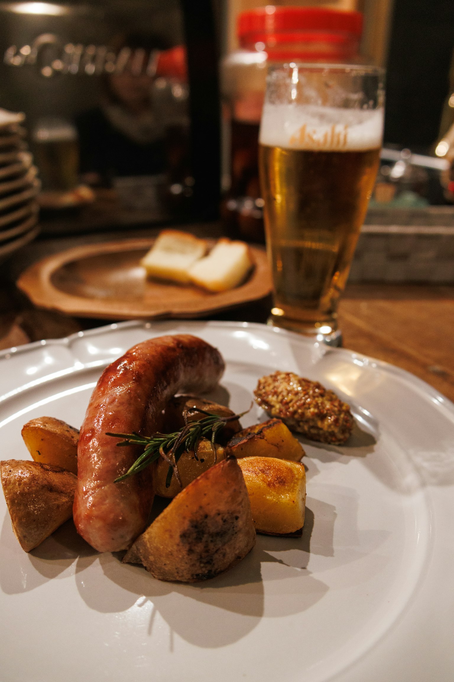 A plate with sausage and potatoes served alongside beer and bread in the background