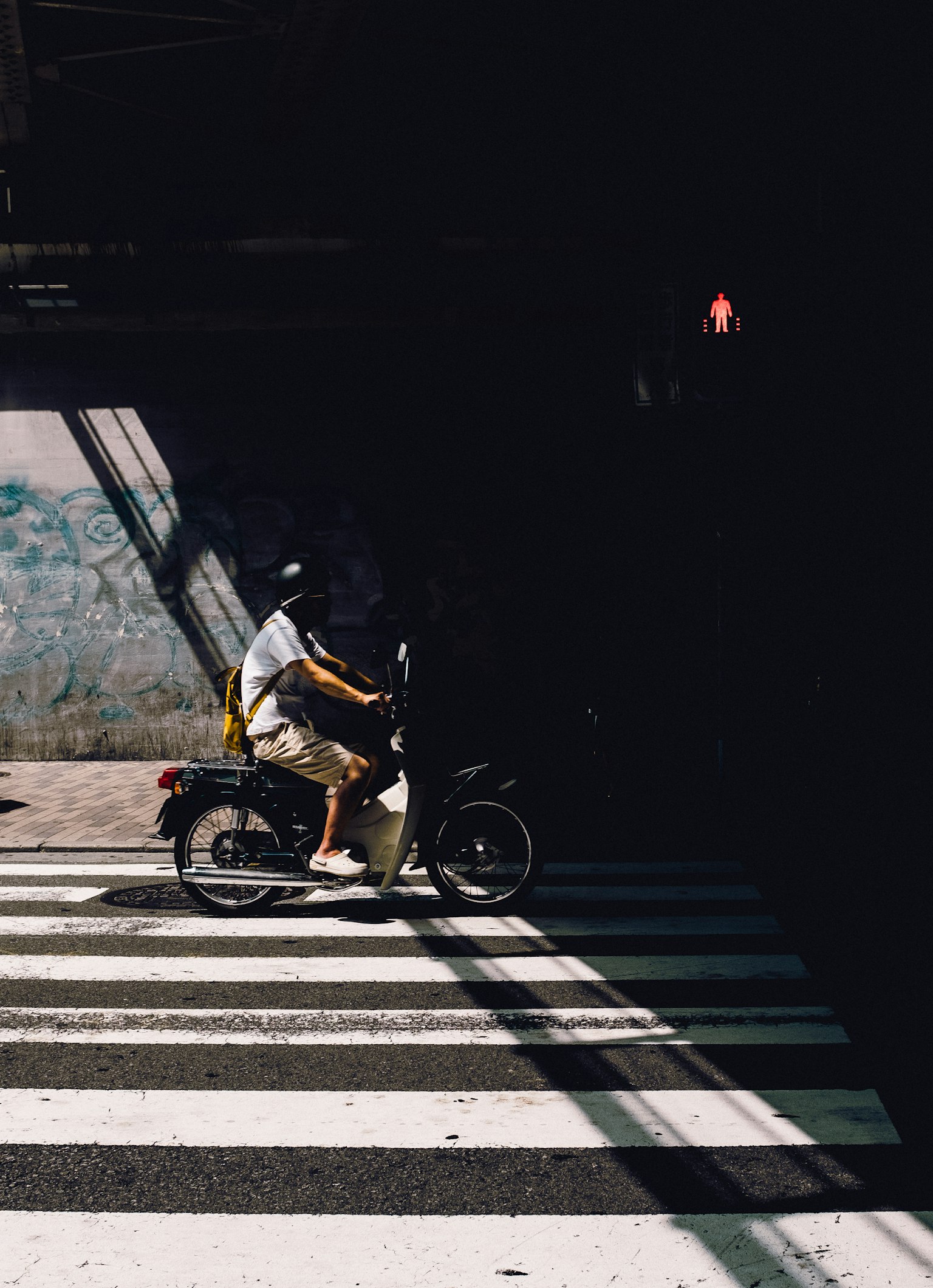A man on a motorcycle crossing a bright crosswalk with contrasting shadows