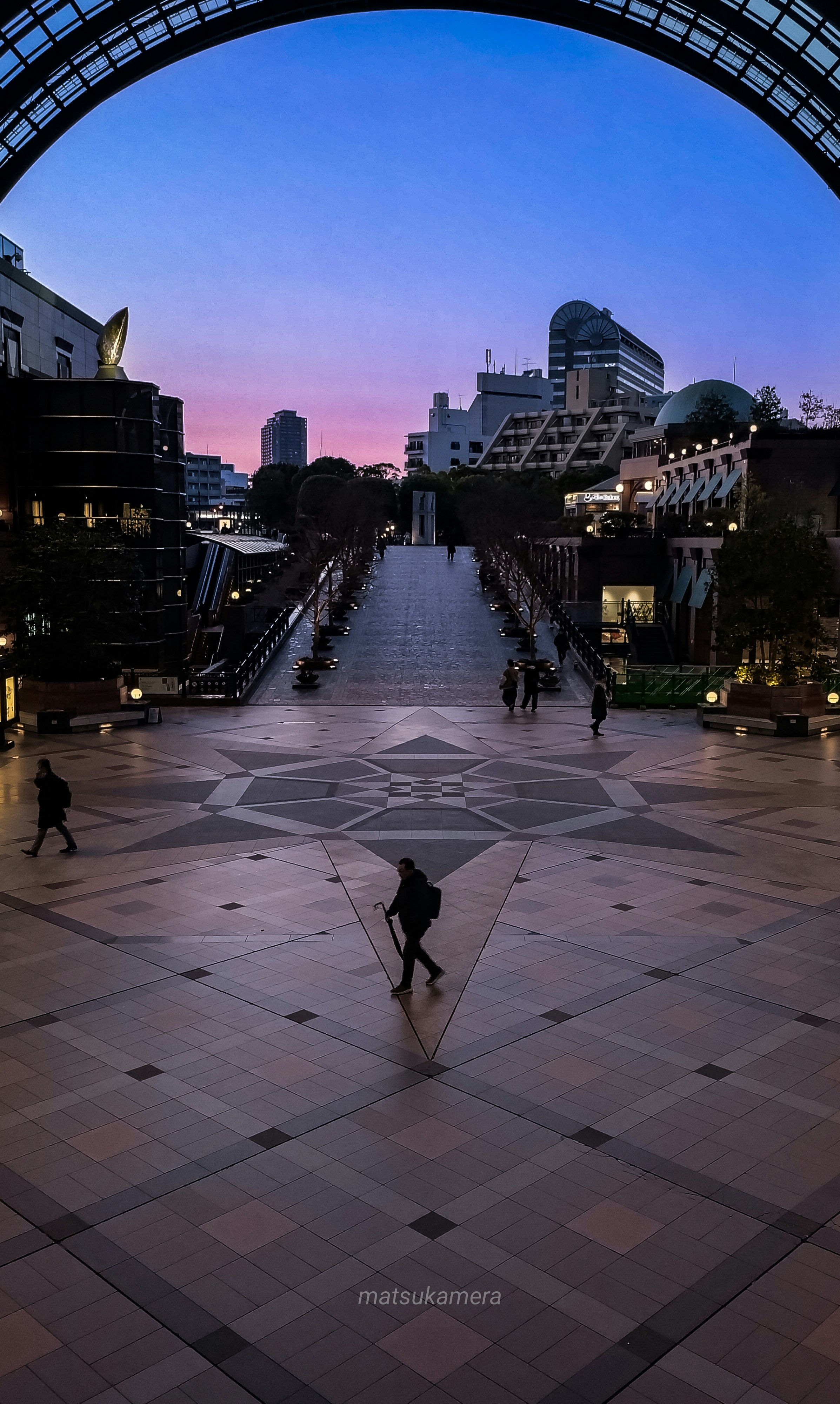 A dusk boulevard and an arched elevated structure in a plaza