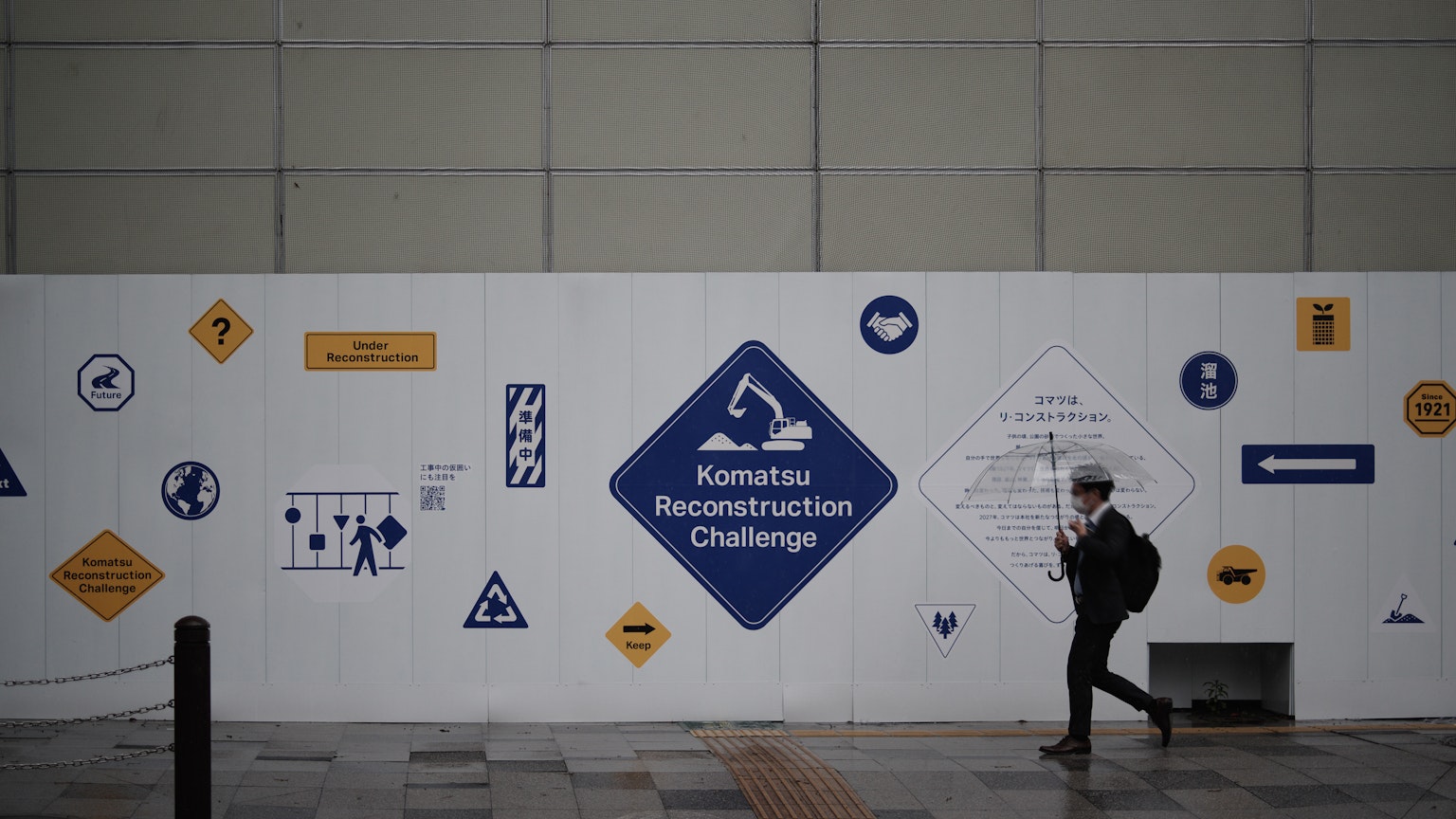 Eine Person, die an einer Wand vorbeigeht, die mit verschiedenen Schildern und Logos dekoriert ist, einschließlich eines auffälligen blauen Diamanten für die 'Rekonstruktionsherausforderung'