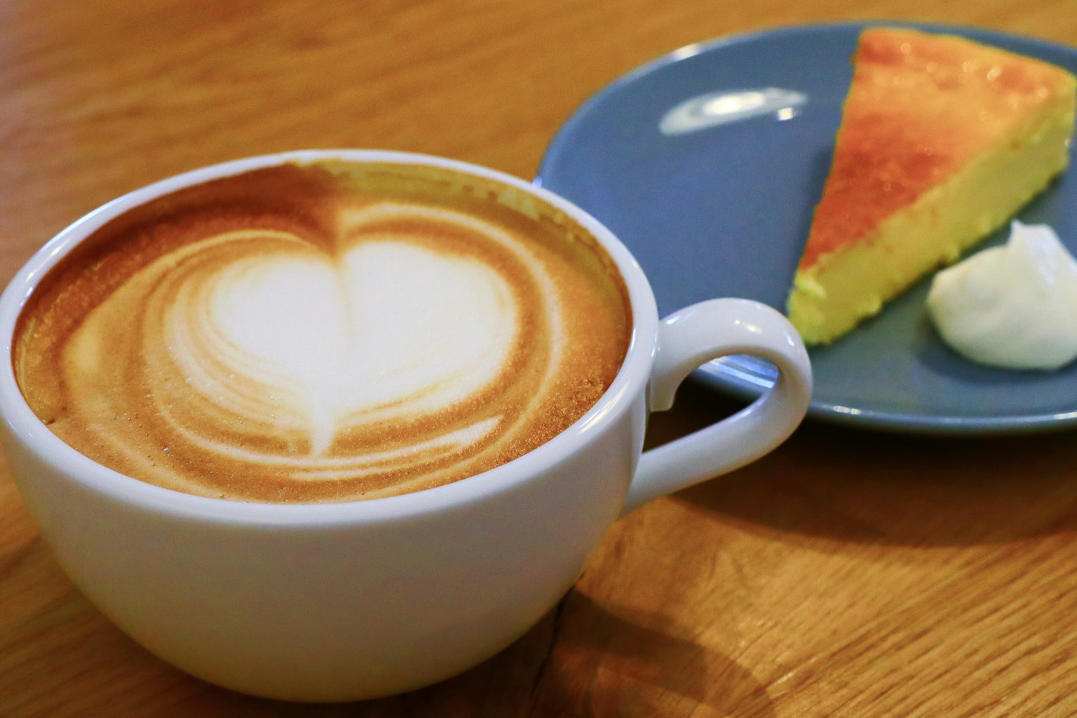 Cà phê với nghệ thuật latte hình trái tim trong cốc trắng kèm theo một lát bánh trên đĩa xanh