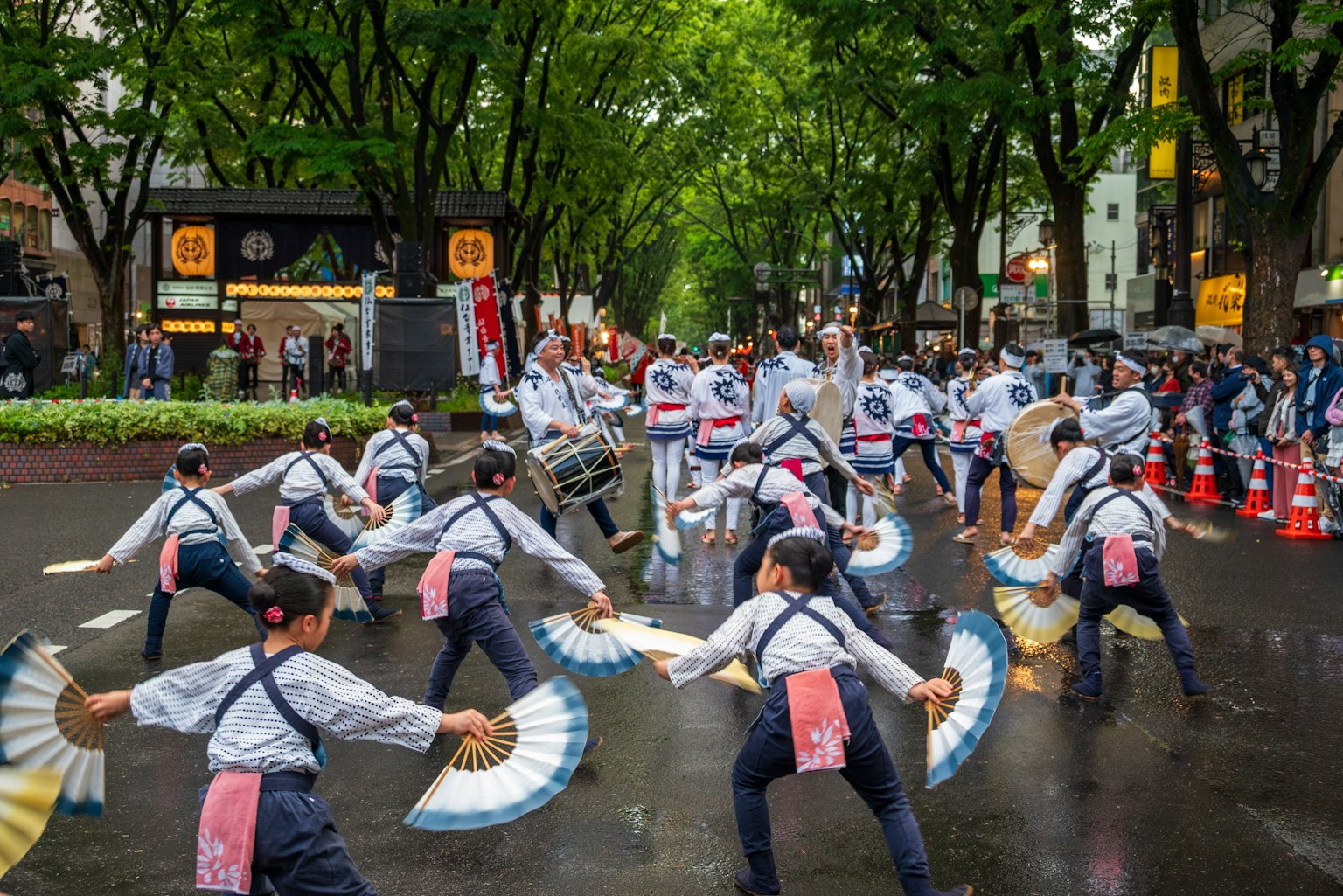ผู้เข้าร่วมเทศกาลญี่ปุ่นแบบดั้งเดิมเต้นรำด้วยพัดบนถนนที่มีฝนตกและมีต้นไม้เรียงราย