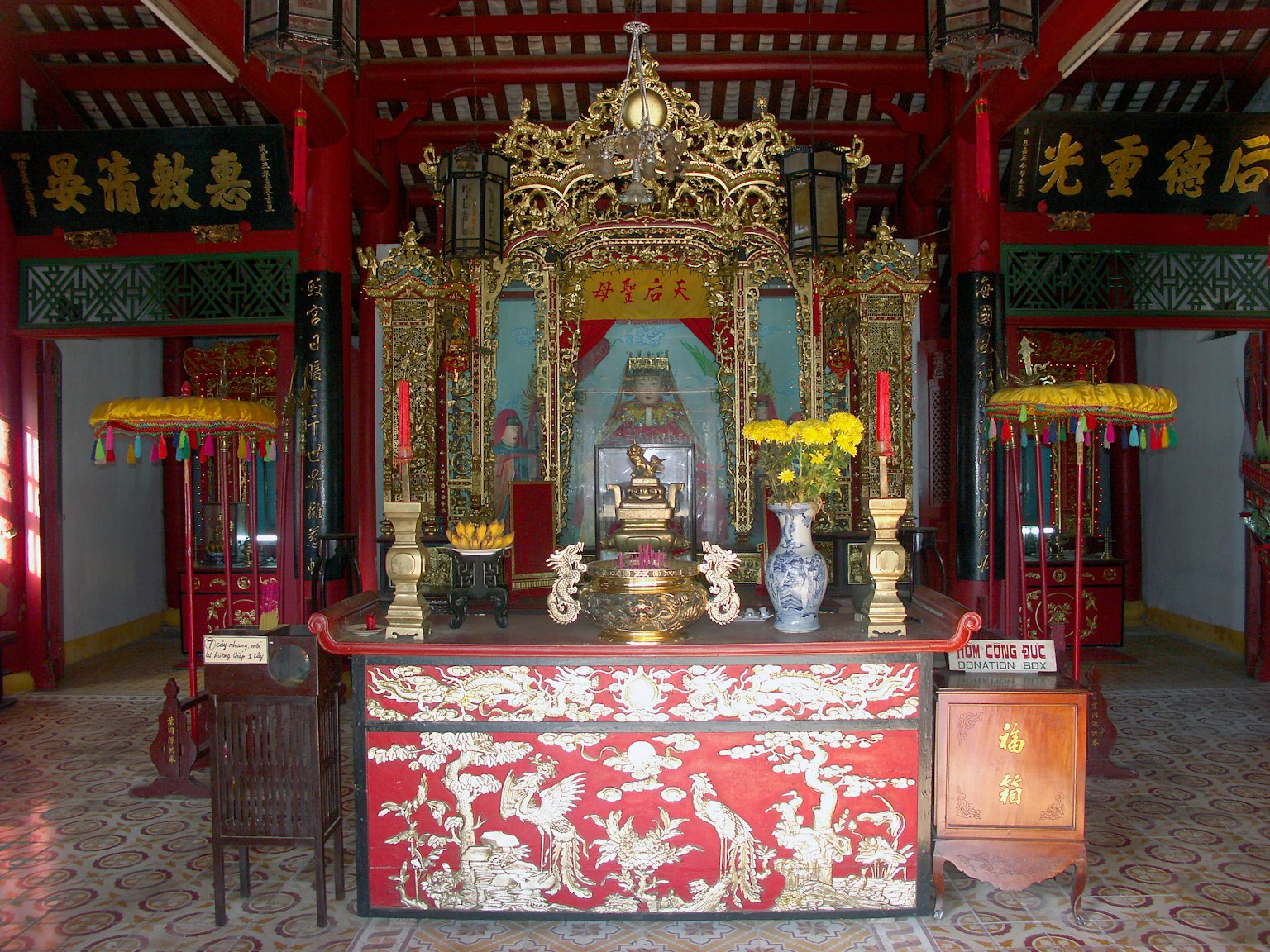 Interior view of a traditional temple with a red and gold altar featuring a statue in the center surrounded by vases and lanterns