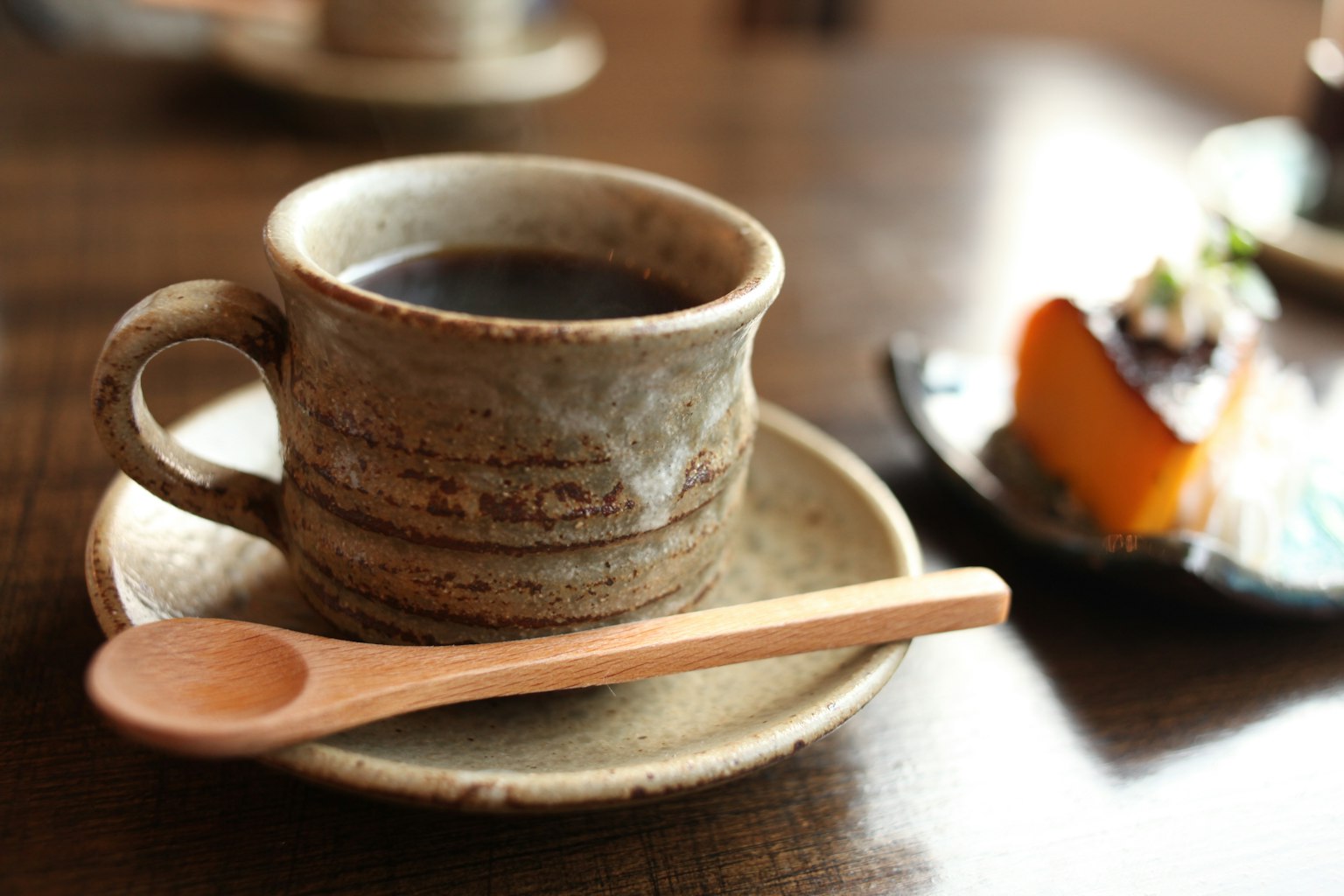Keramiktasse mit Kaffee und Holzlöffel sowie Dessert im Hintergrund