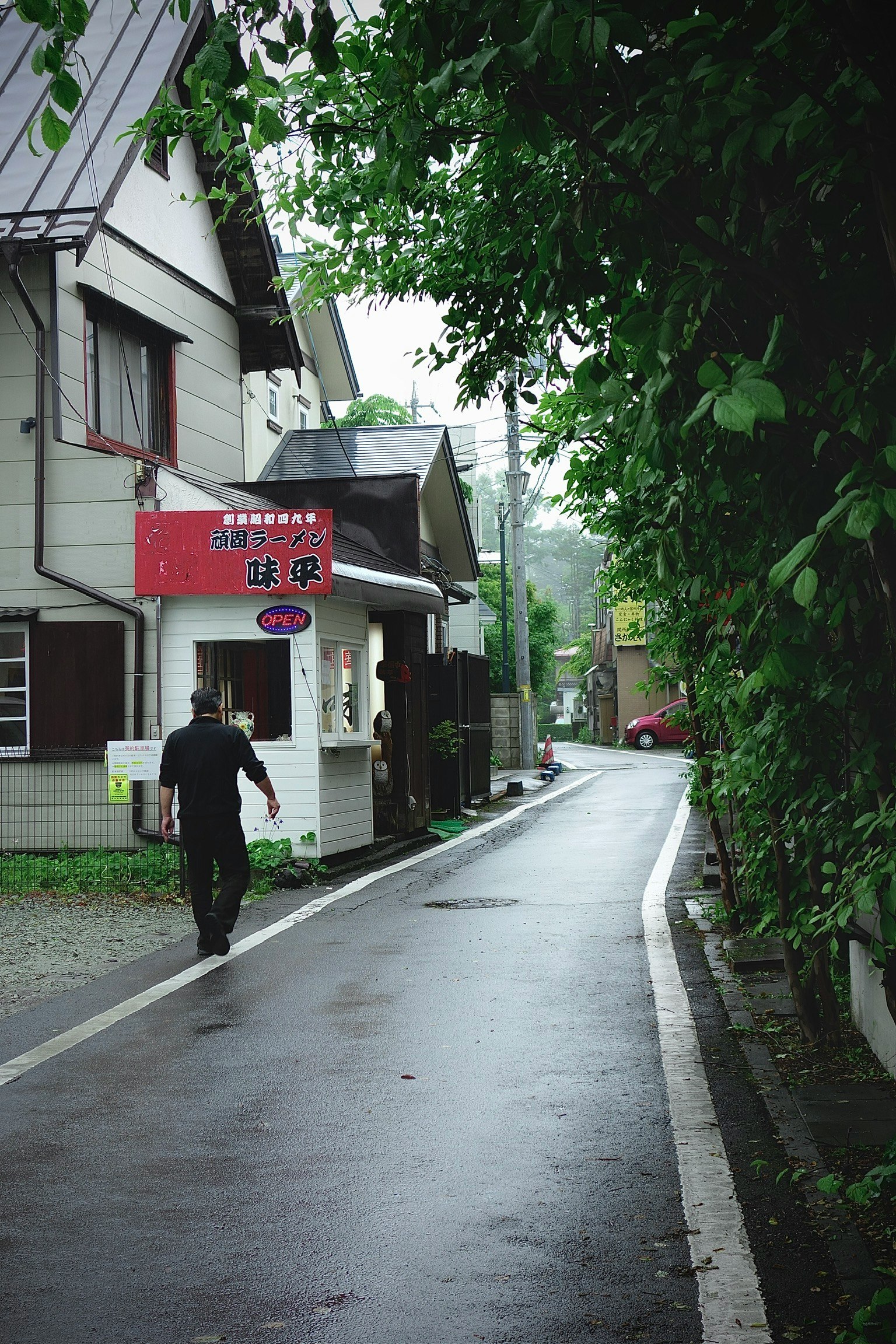 一個人在雨天走在狹窄的道路上，周圍是綠色的樹木和房子