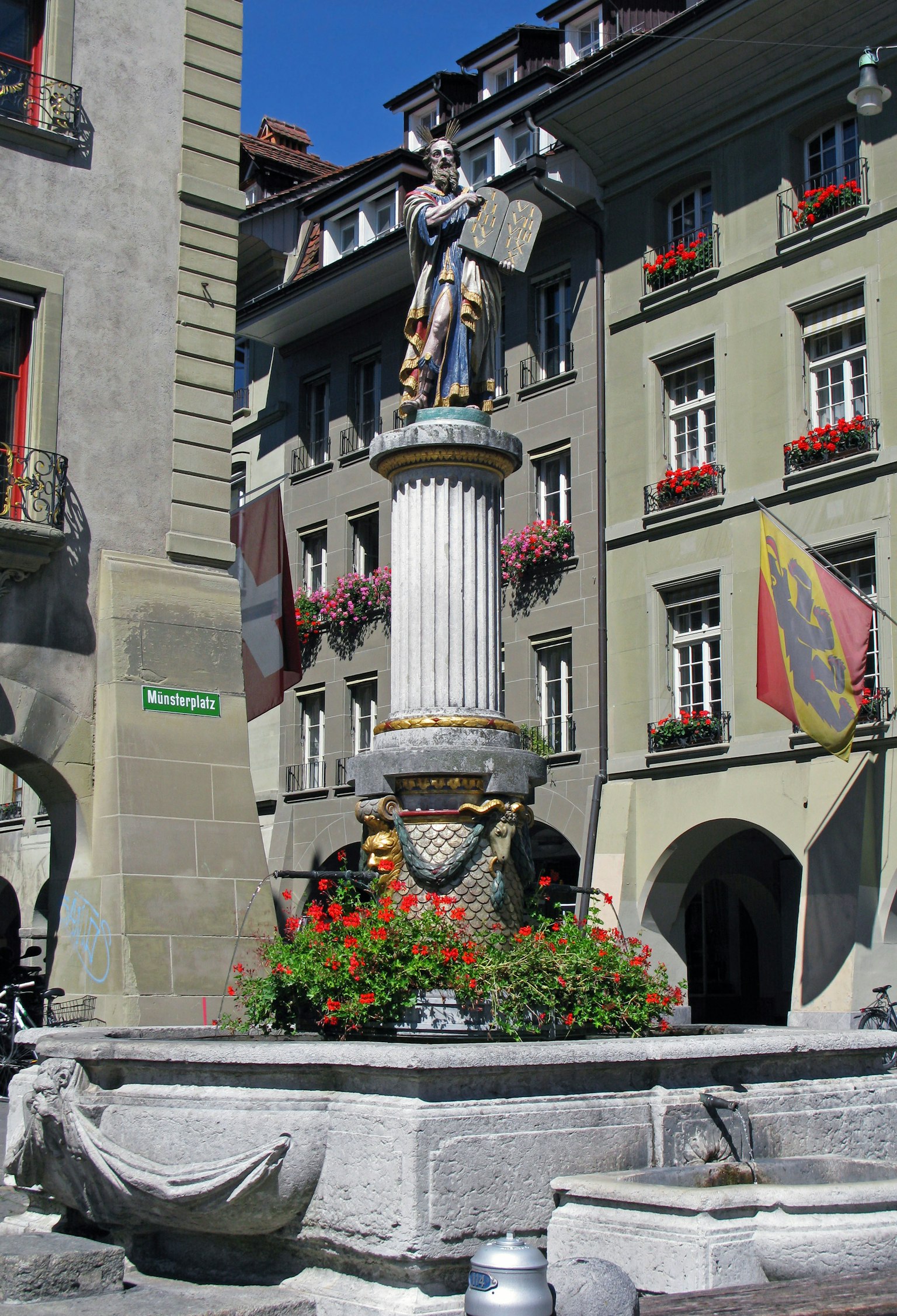Une place avec une fontaine et une statue sur une colonne entourée de bâtiments