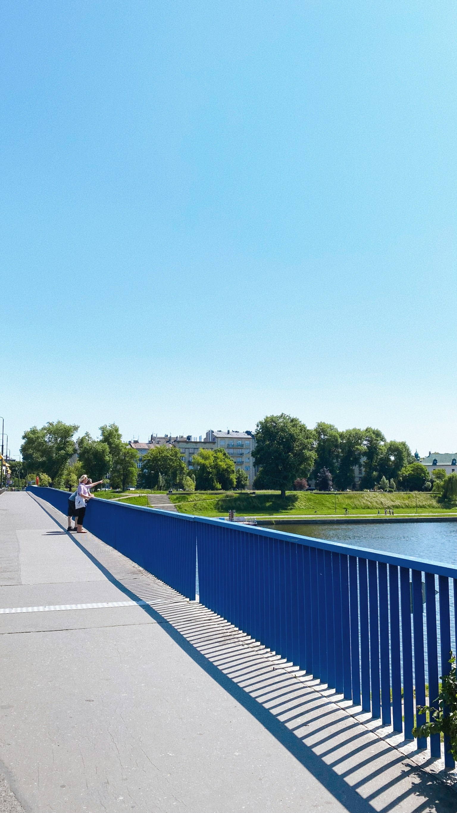 Ponte con parapetto blu che si affaccia su un lago sereno e alberi verdi sotto un cielo azzurro