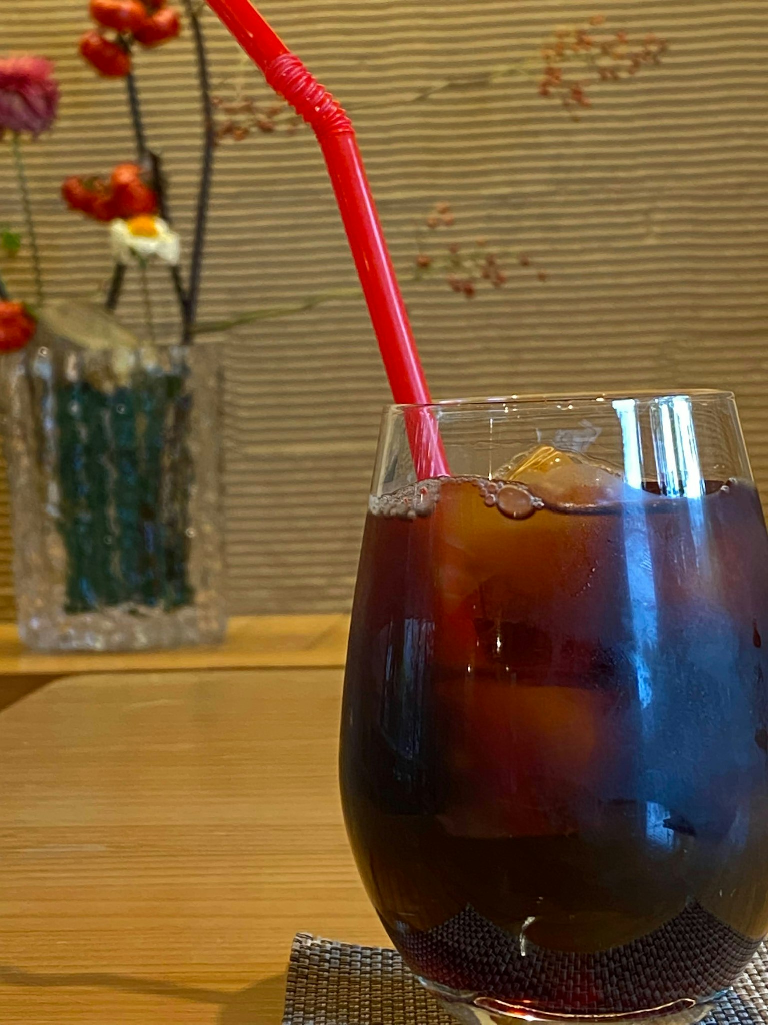Un vaso de bebida helada con una pajita roja y un jarrón de flores al fondo