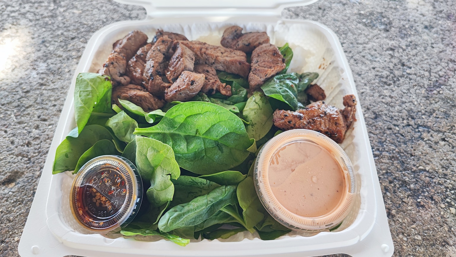 Kotak makanan dengan daging sapi panggang di atas salad bayam dan dua saus