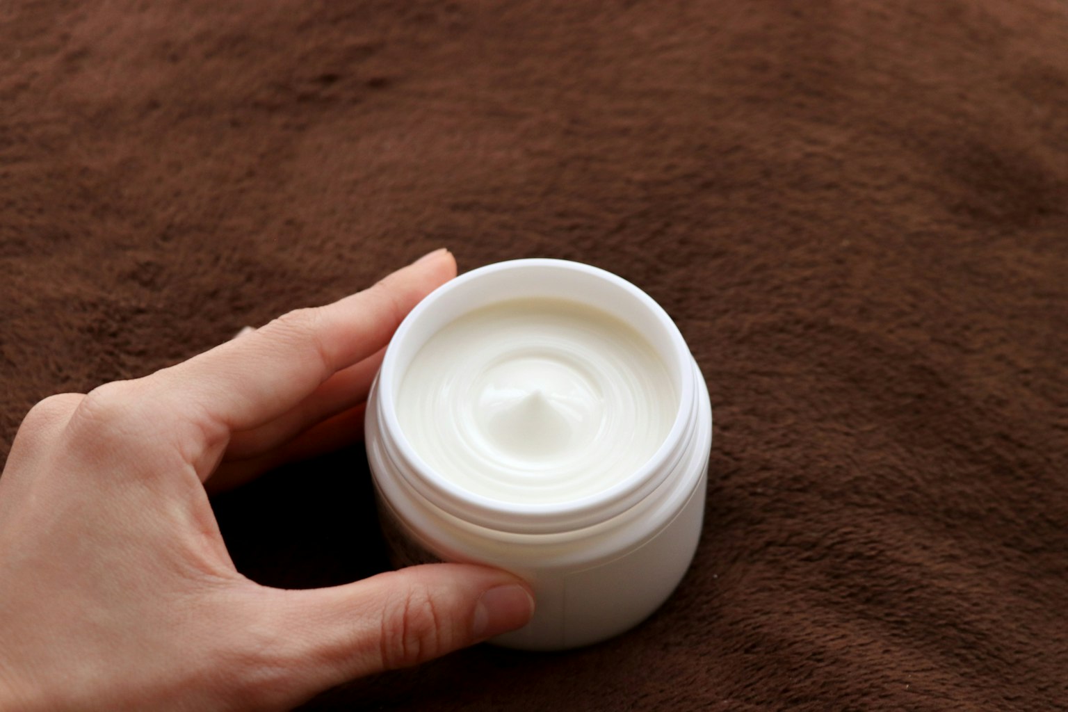 Une main tenant un pot de crème blanche posé sur un tissu marron