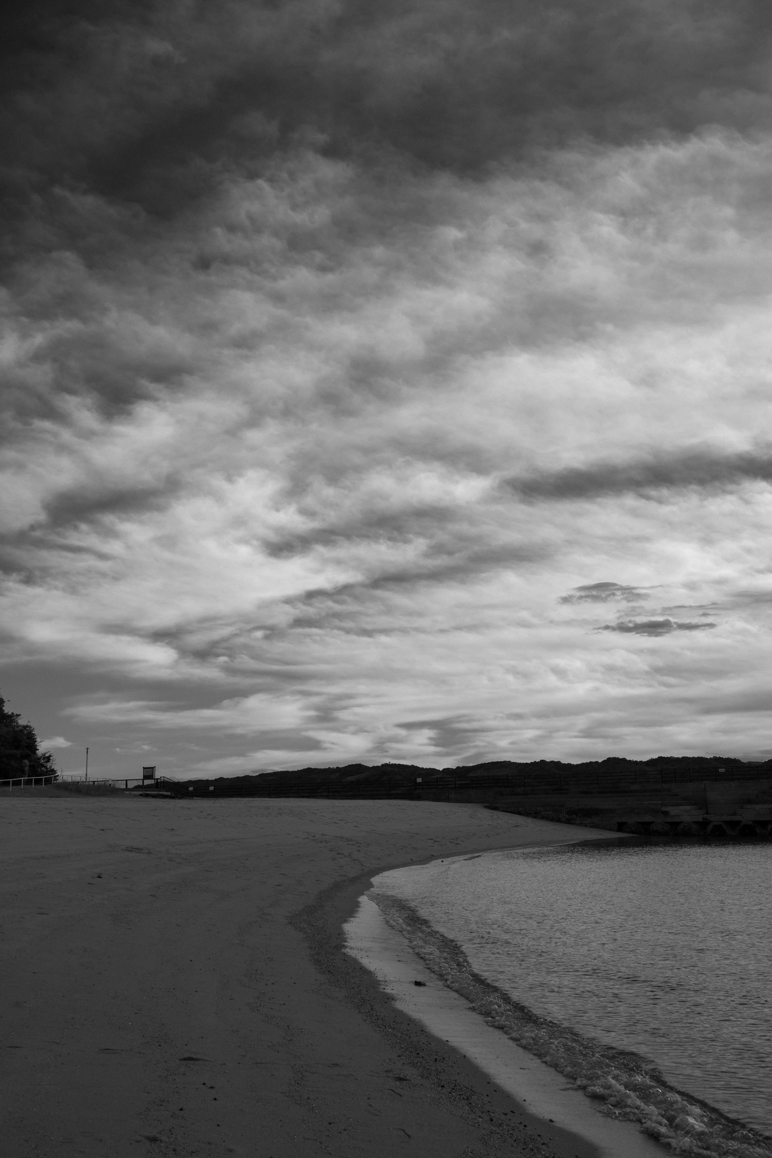 Pemandangan pantai hitam putih dengan gelombang tenang yang menghantam pasir dan langit mendung