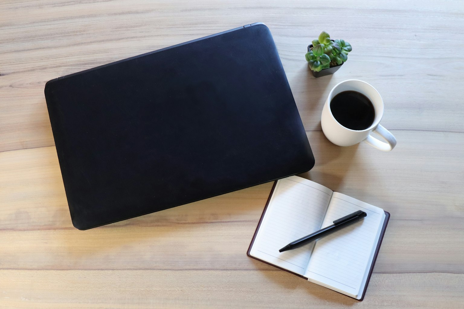 Schwarzes Laptop auf einem Holzschreibtisch mit einer weißen Kaffeetasse einem Notizbuch und einem Stift