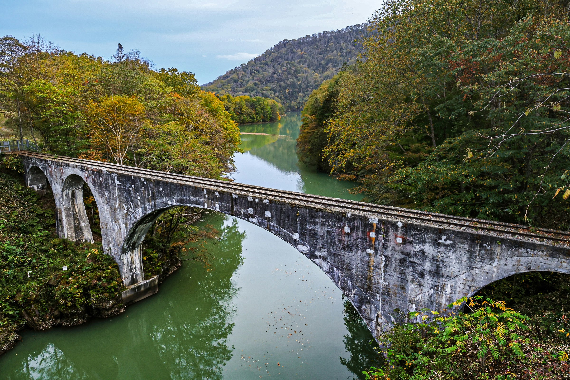 秋の渓谷を流れる緑の川と石造りのアーチ橋の風景