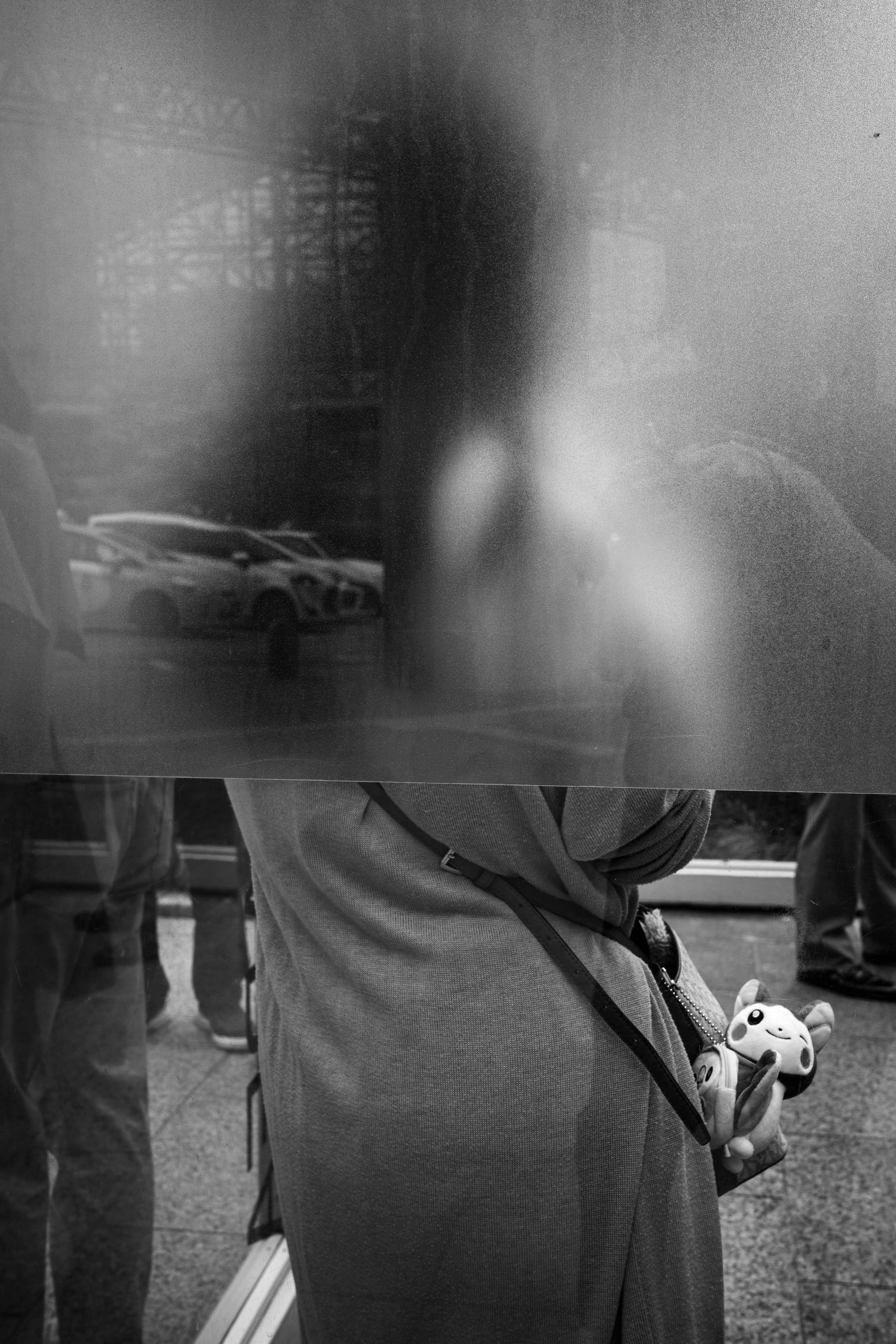 ガラス越しのモノクロ街角で立つ人物とぬいぐるみ